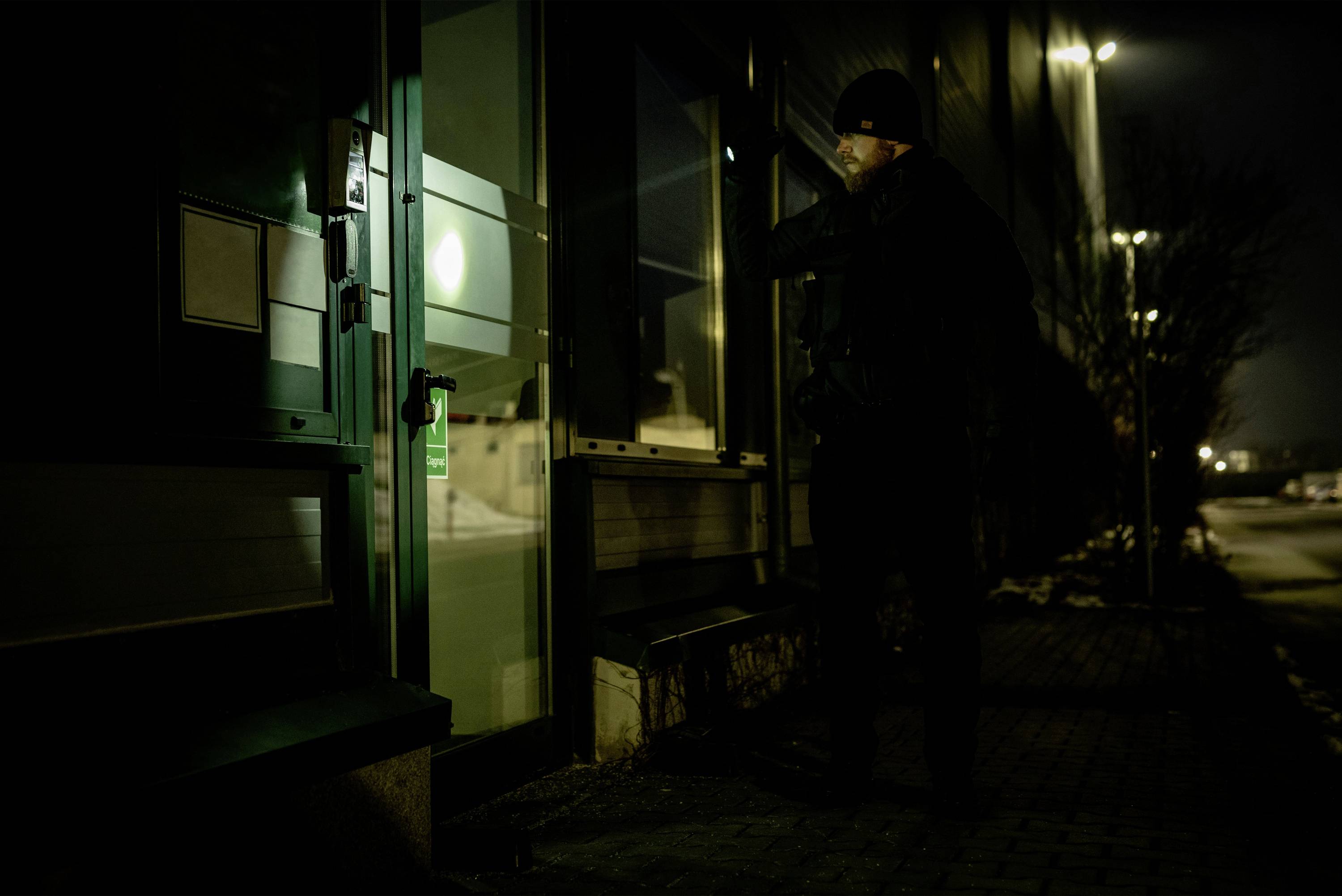 Mężczyzna stoi nocą przed drzwiami wejściowymi i świeci latarką. Otoczenie ciemne, atmosfera tajemnicza.