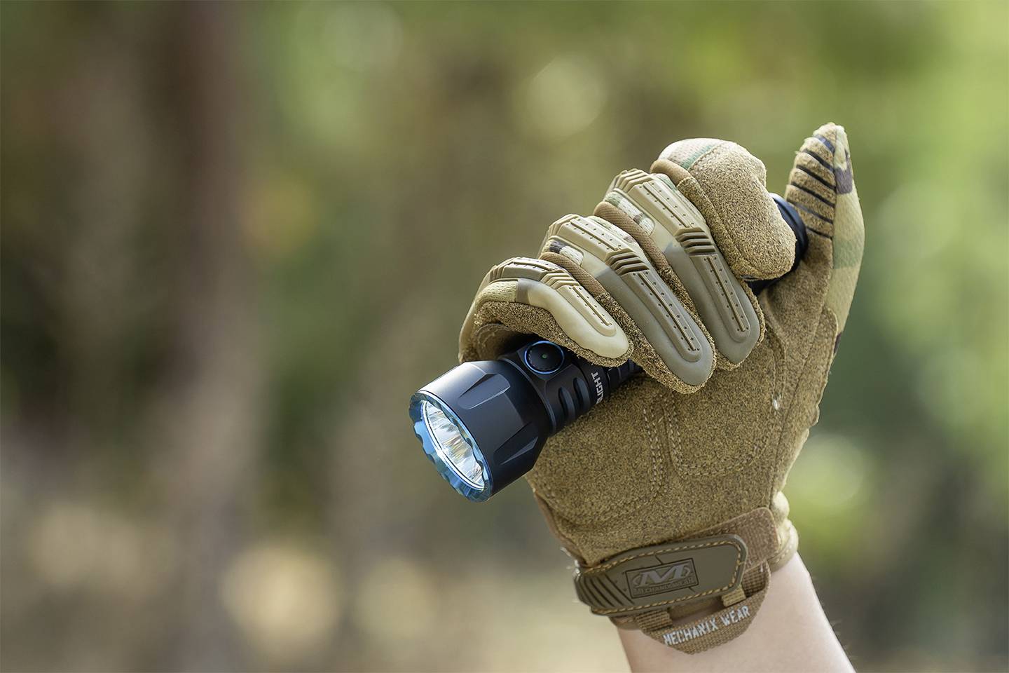 Dłoń w brązowej rękawiczce trzyma włączoną latarkę na wolnym powietrzu. Tło jest rozmyte.