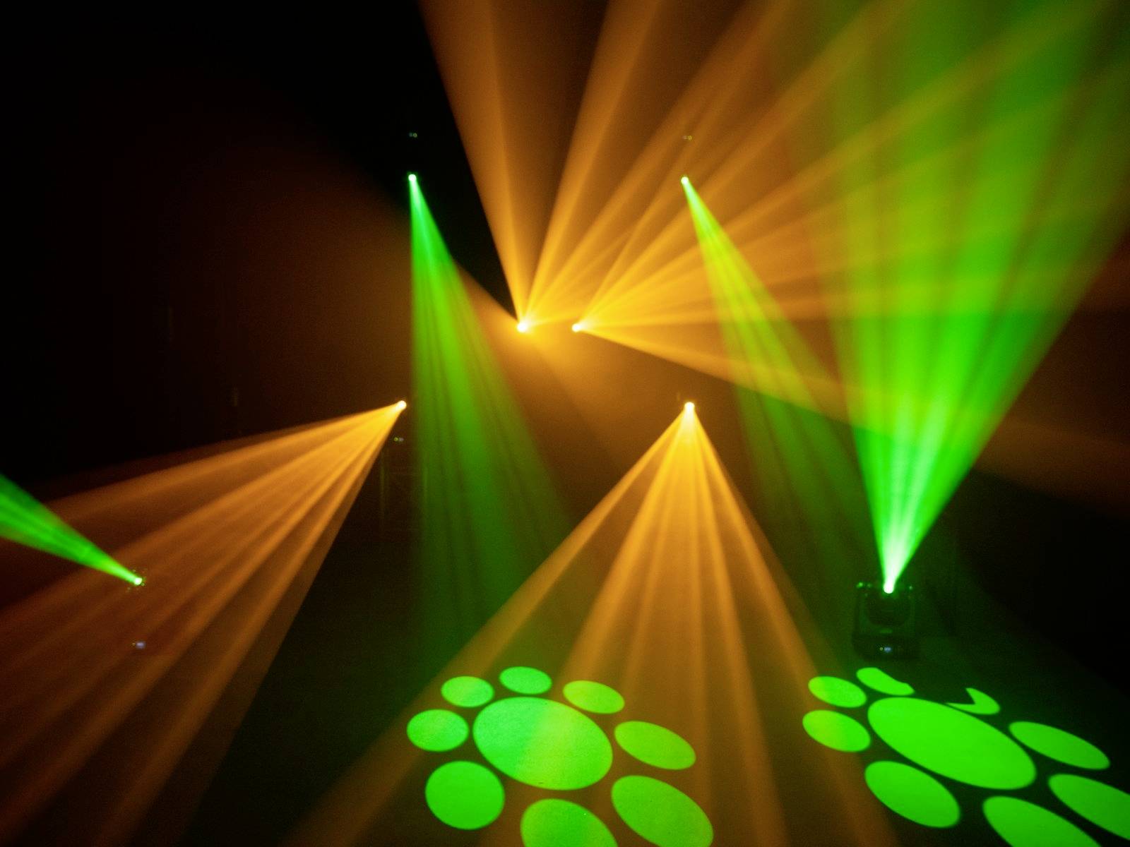 Scena z kolorowymi efektami świetlnymi, zielone i żółte promienie świecą w ciemnym pomieszczeniu. Na podłodze widoczne są okrągłe wzory.