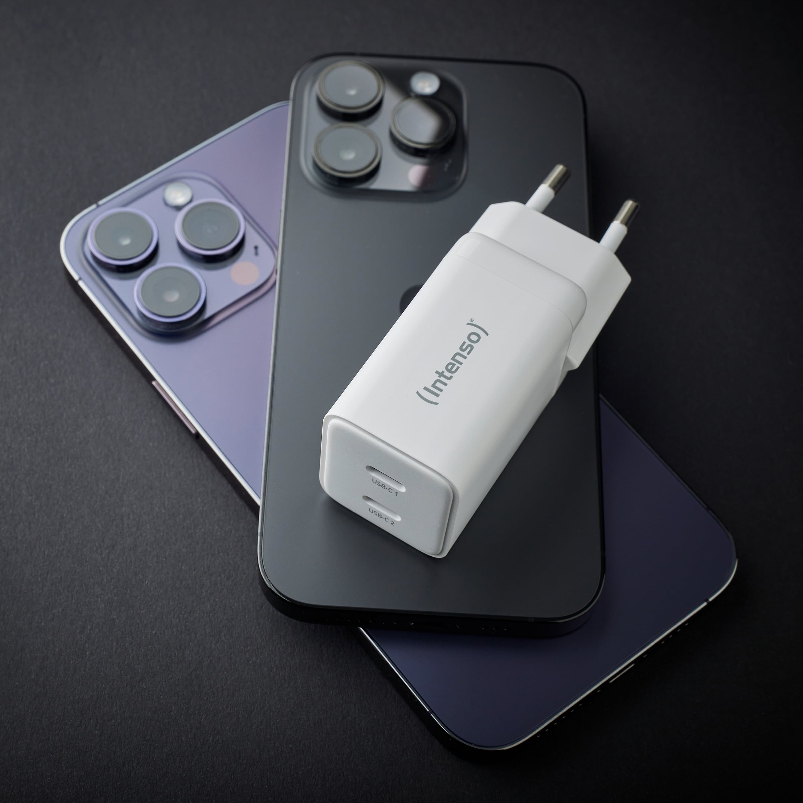 Dwa smartfony z trzema obiektywami aparatów, jeden w kolorze fioletowym, a drugi w czerni, leżą na sobie. Biała ładowarka spoczywa na wierzchu.
