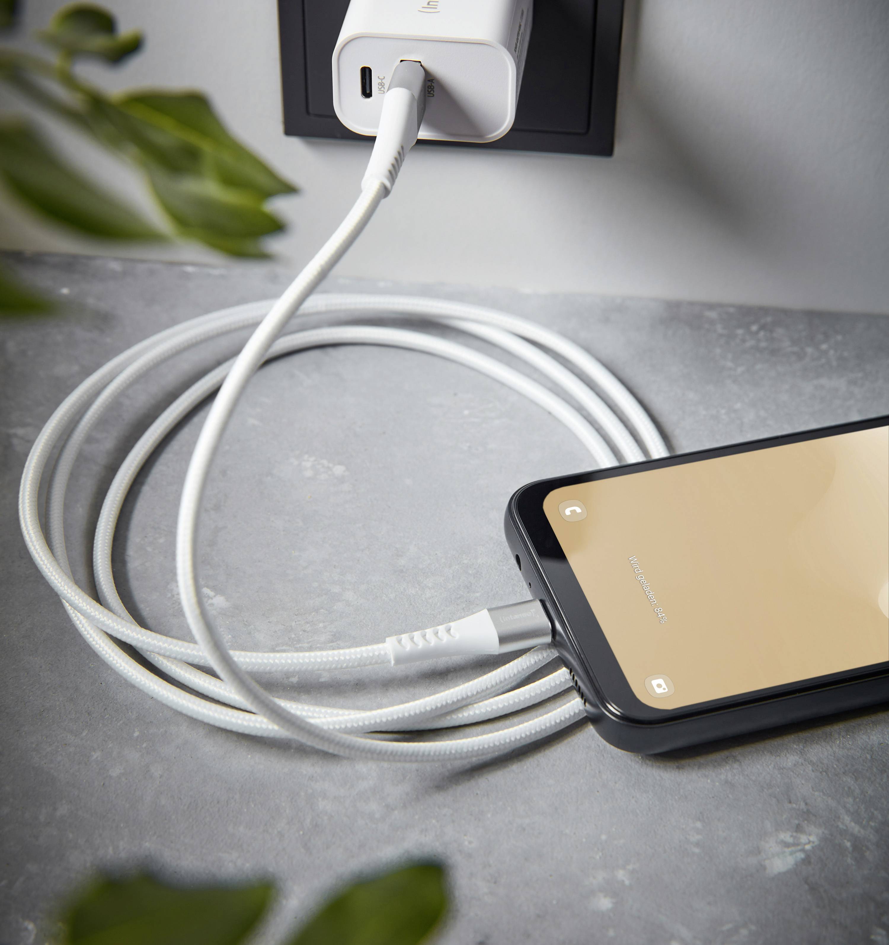 Smartfon jestładowany białym kablem przy gniazdku elektrycznym. Telefon leży na stole, a kabel jest zwinięty.