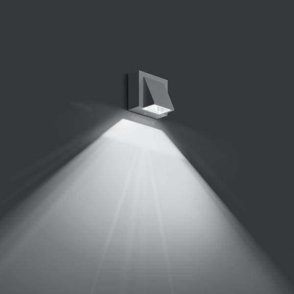 Nowoczesna lampa ścienna świeci w ciemnym pomieszczeniu. Rzuca jasne, równomierne światło pod szerokim kątem w dół.