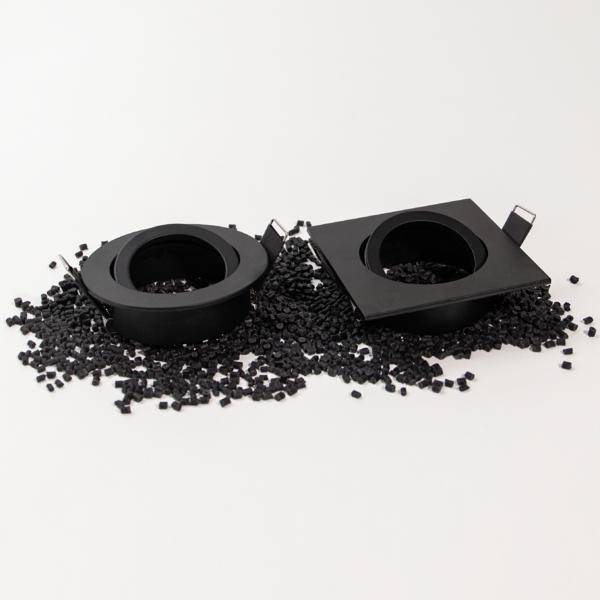 Dwa czarne plastikowe elementy - jeden w kształcie koła, drugi w kształcie kwadratu - leżą na stosie czarnych granulek plastikowych na białym tle.