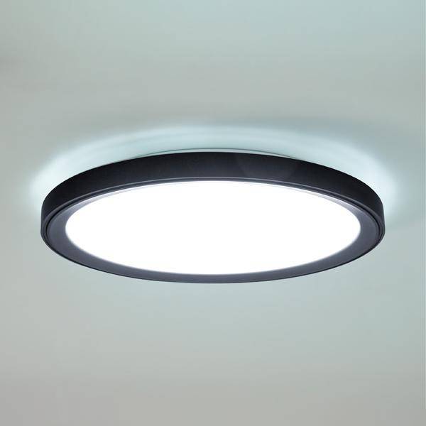 Okrągła, płaska lampa sufitowa z czarną obwódką świeci jasno. Znajduje się w minimalistycznie urządzonym pomieszczeniu.