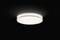 Okrągła lampa sufitowa z rozproszonym, białym światłem w ciemnym pomieszczeniu. Lampa jest umieszczona centralnie na zdjęciu i włączona.