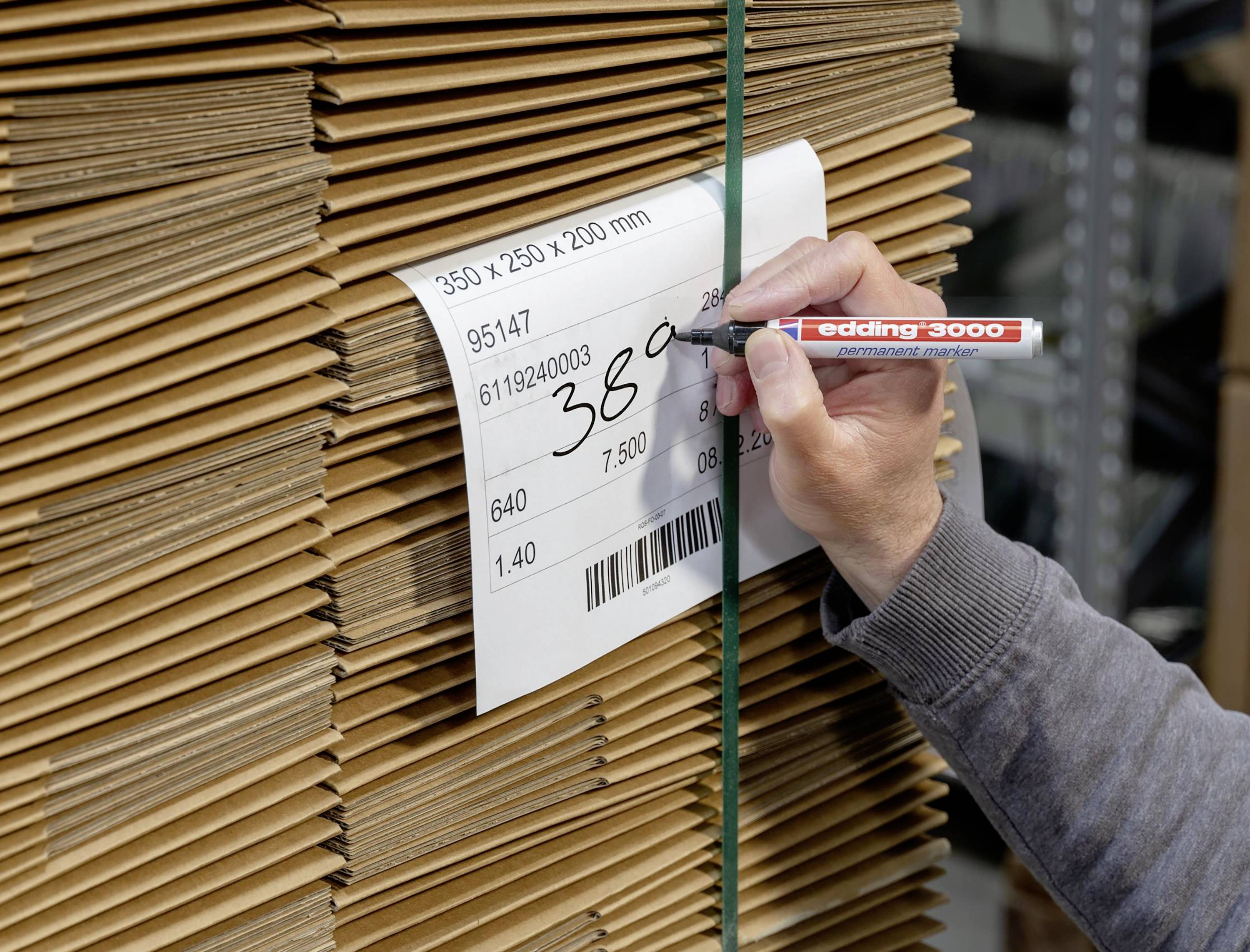 Stos kartonów z etykietą jest opisywany przez osobę piszącą długopisem. Fokus skierowany jest na tekst etykiety.