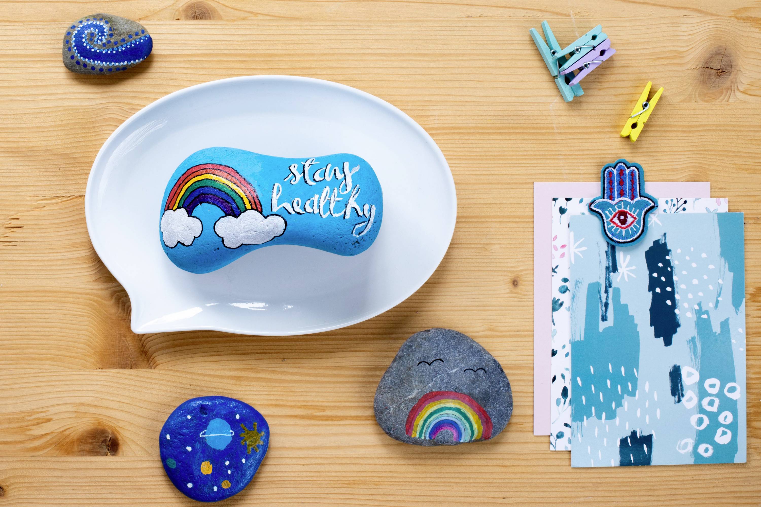 Dekoracyjny stolik z malowanymi kamieniami, jeden z napisem 'stay healthy', obok kolorowe wzorniki i małe spinacze.