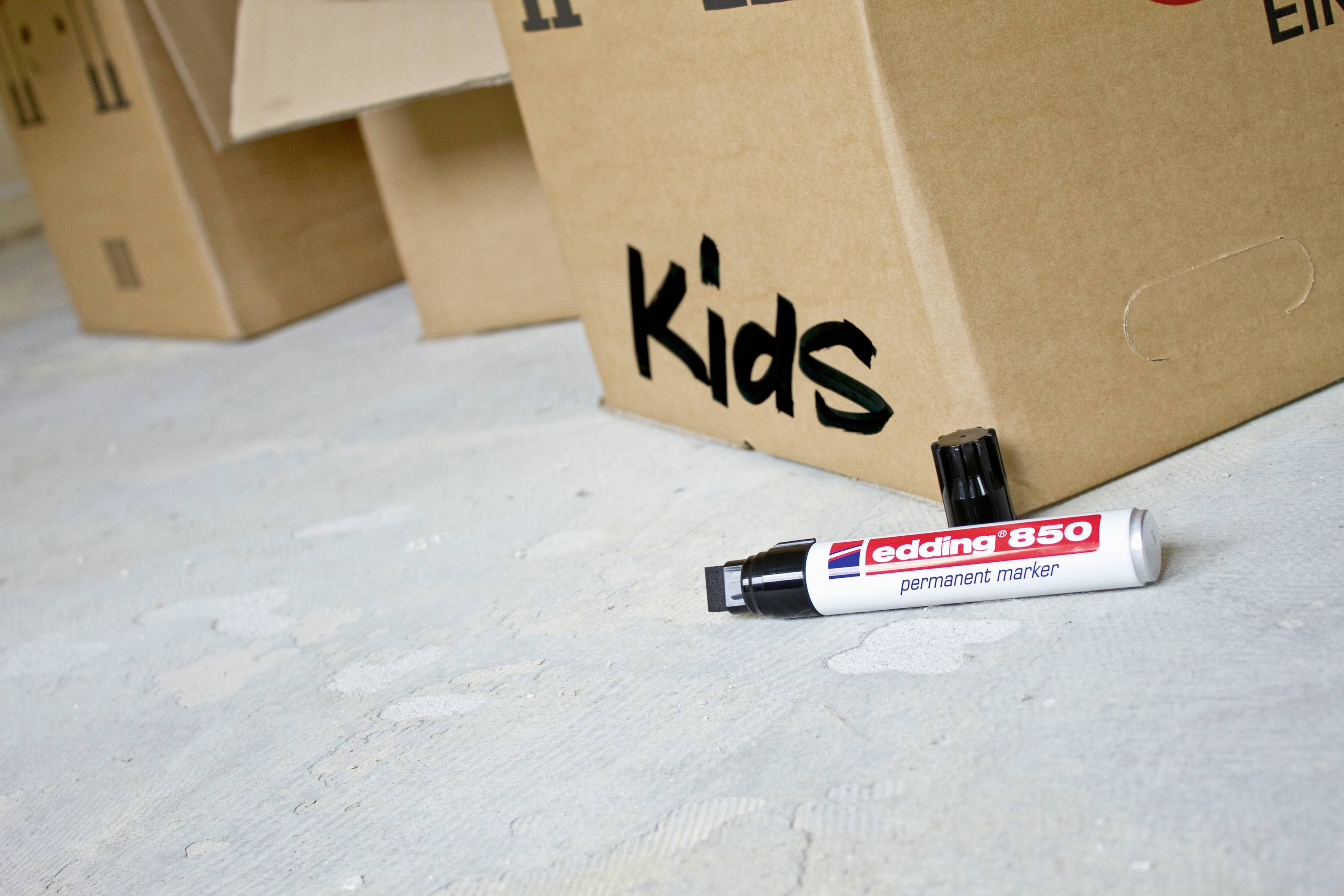 Kartony na podłodze, jeden z nich opisany napisem 'Kids'. Przed nimi leży marker permanentny Edding 850.