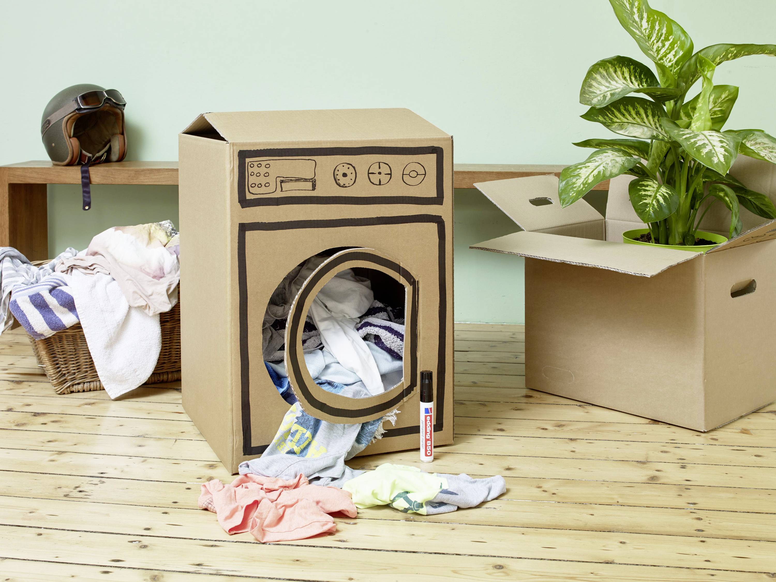 Wykonany z kartonu zabawkowy suszar do ubrań stoi na drewnianej podłodze. Obok niego leżą i wiszą kolorowe ubrania.
