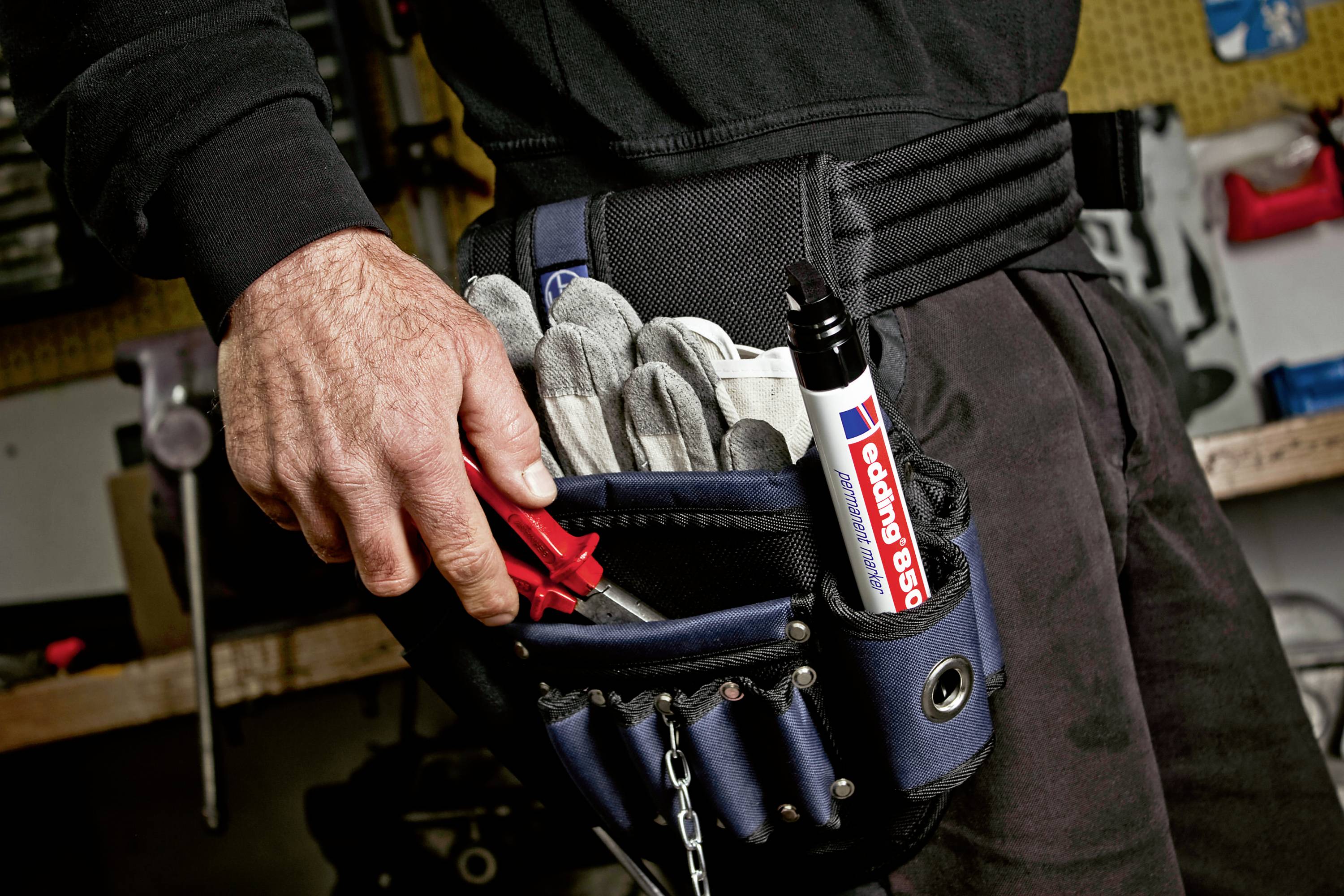 Rzemieślnik trzyma w ręku czerwone szczypce. W czarnym pasie narzędziowym umieszczone są rękawice robocze i duży marker.