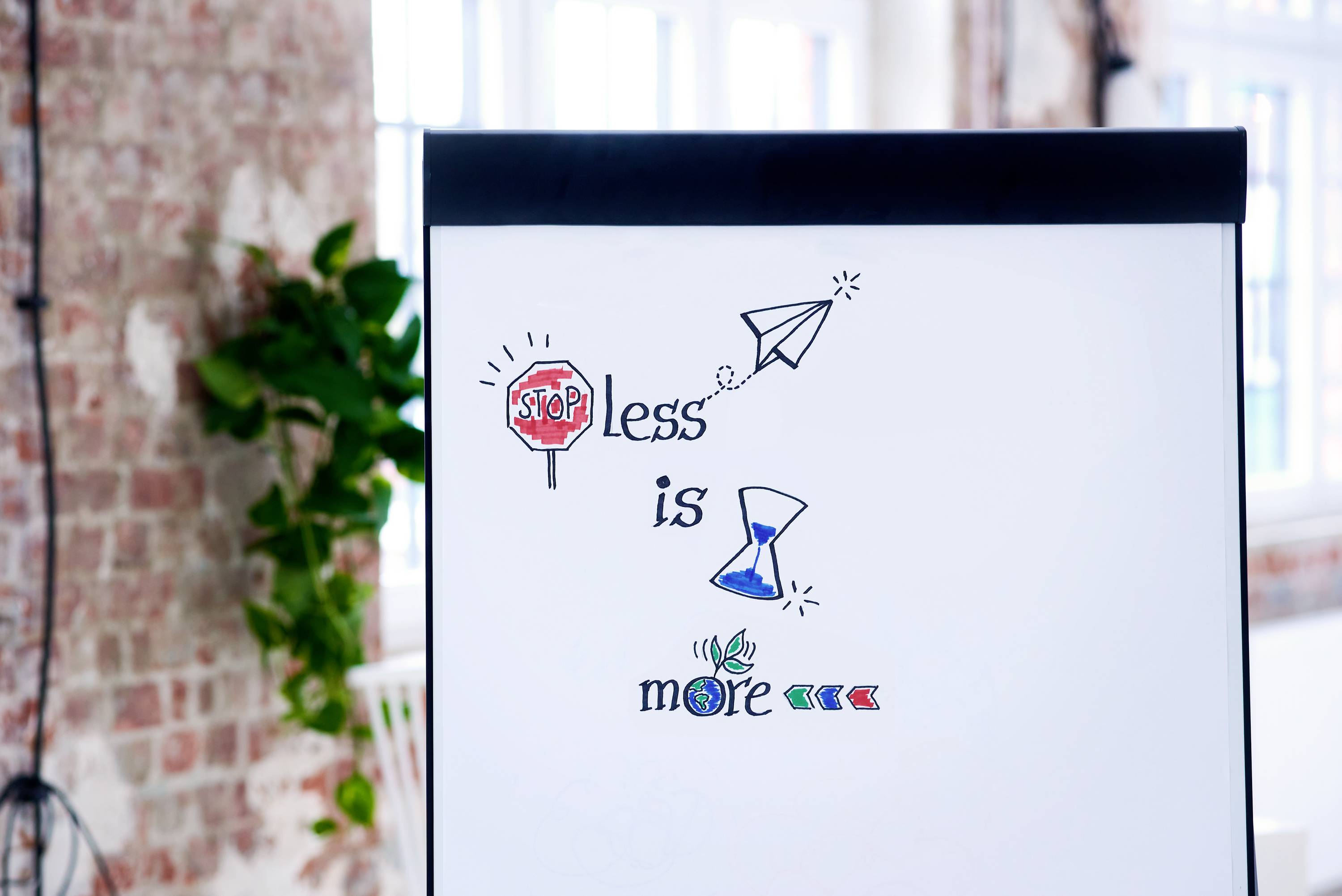 Flipchart prezentuje frazę 'Mniej znaczy więcej', zilustrowaną znakiem stopu, papierowym samolotem, klepsydrą oraz symbolem wzrostu.