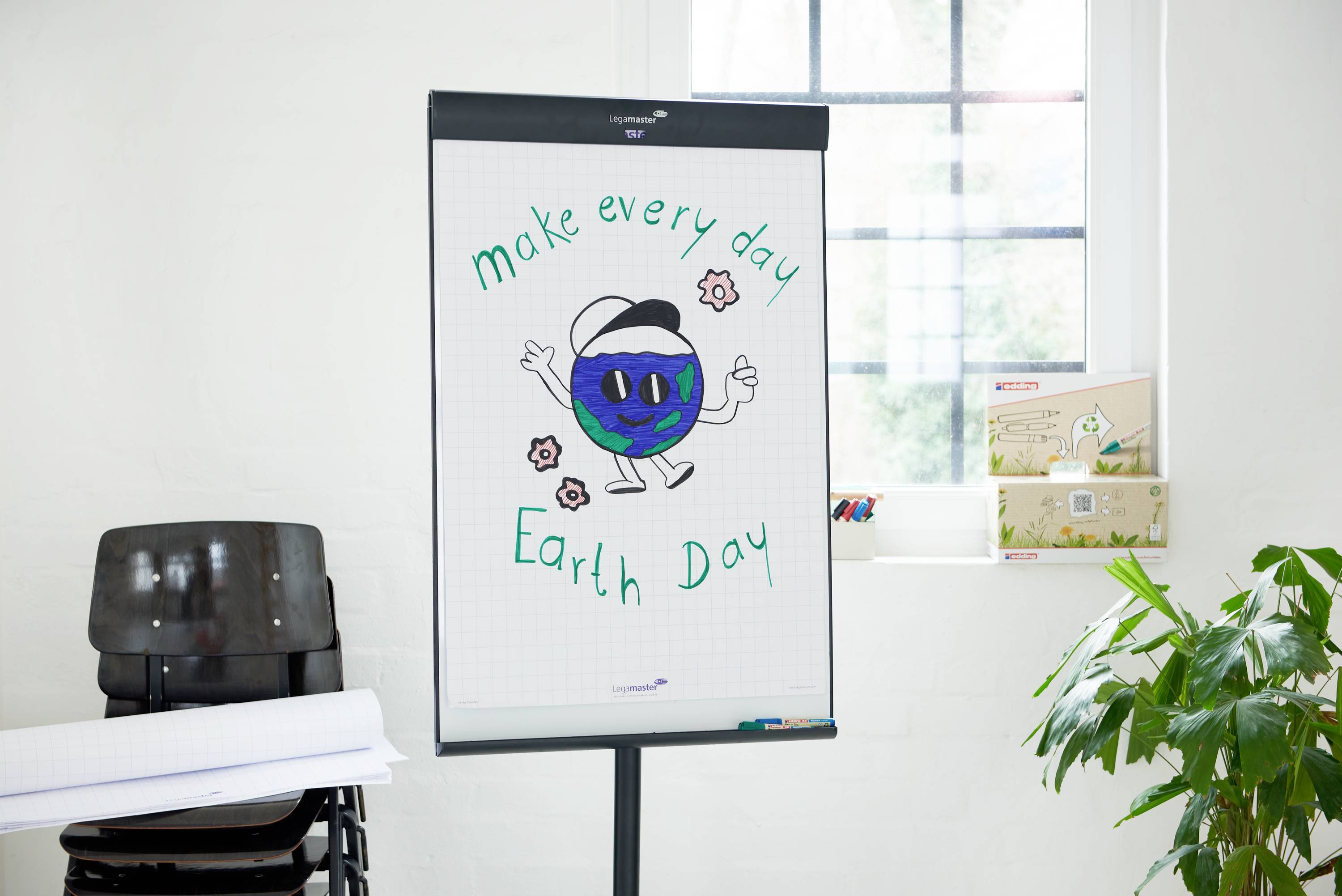 Tablica flipchart z rysunkiem uśmiechniętej planety z rękami i nogami, otoczonej kwiatami i tekstem 'make every day Earth Day'. Pomieszczenie z oknami i roślinami w tle.