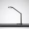 Nowoczesna, czarna lampa biurkowa z przegubowym ramieniem stoi na ciemnym stole.