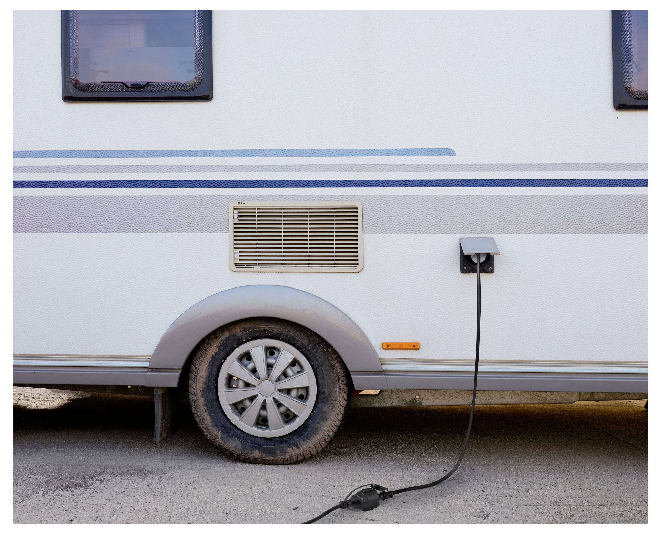Samochód kempingowy zaparkowany na gruntowej drodze z podłączonym wtyczką elektryczną do jego boku, blisko koła, wskazując na ładowanie.
