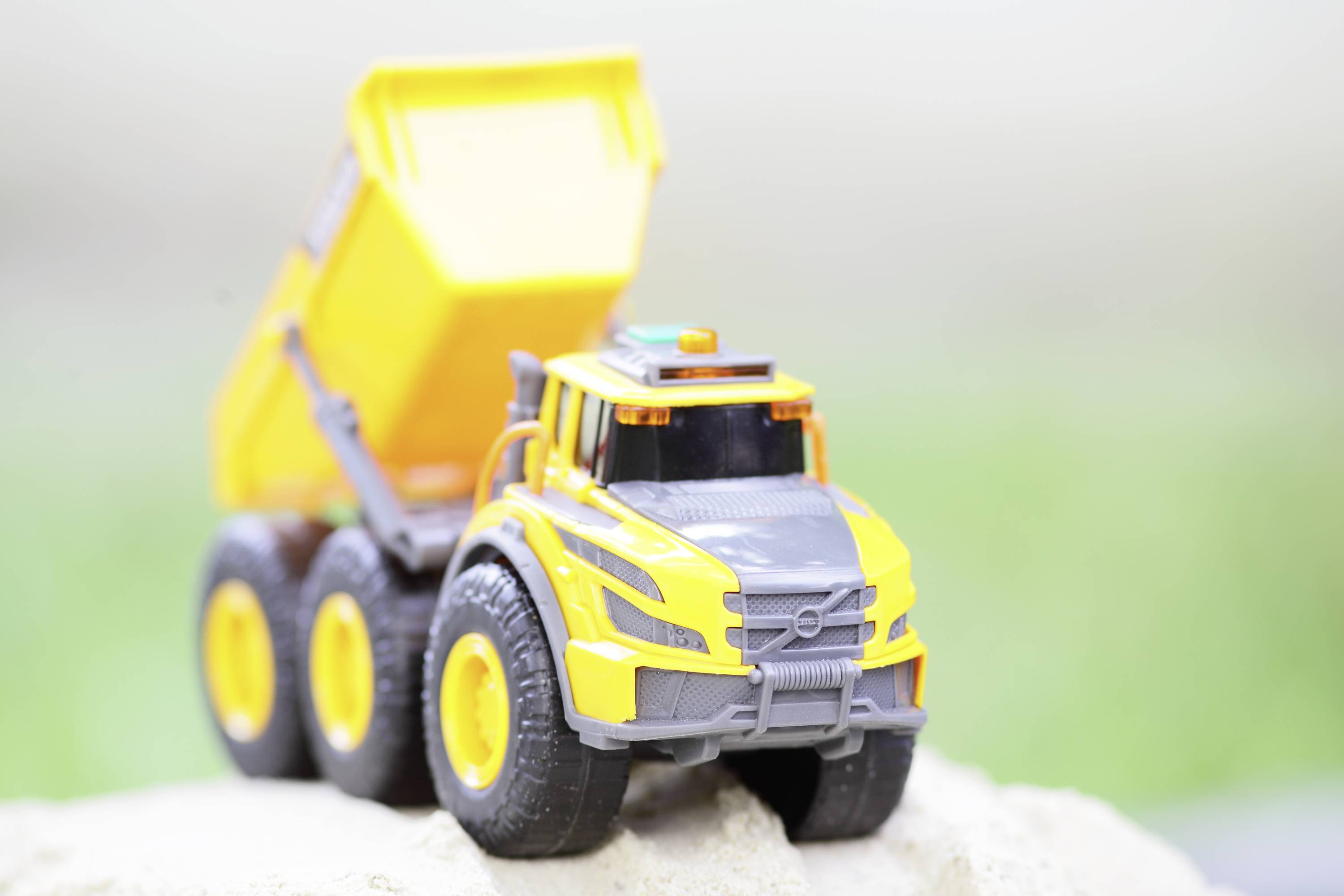 Żółta zabawkowa wywrotka na piaszczystym podłożu. Wywrotka ma uniesioną skrzynię ładunkową i posiada sześć dużych kół.