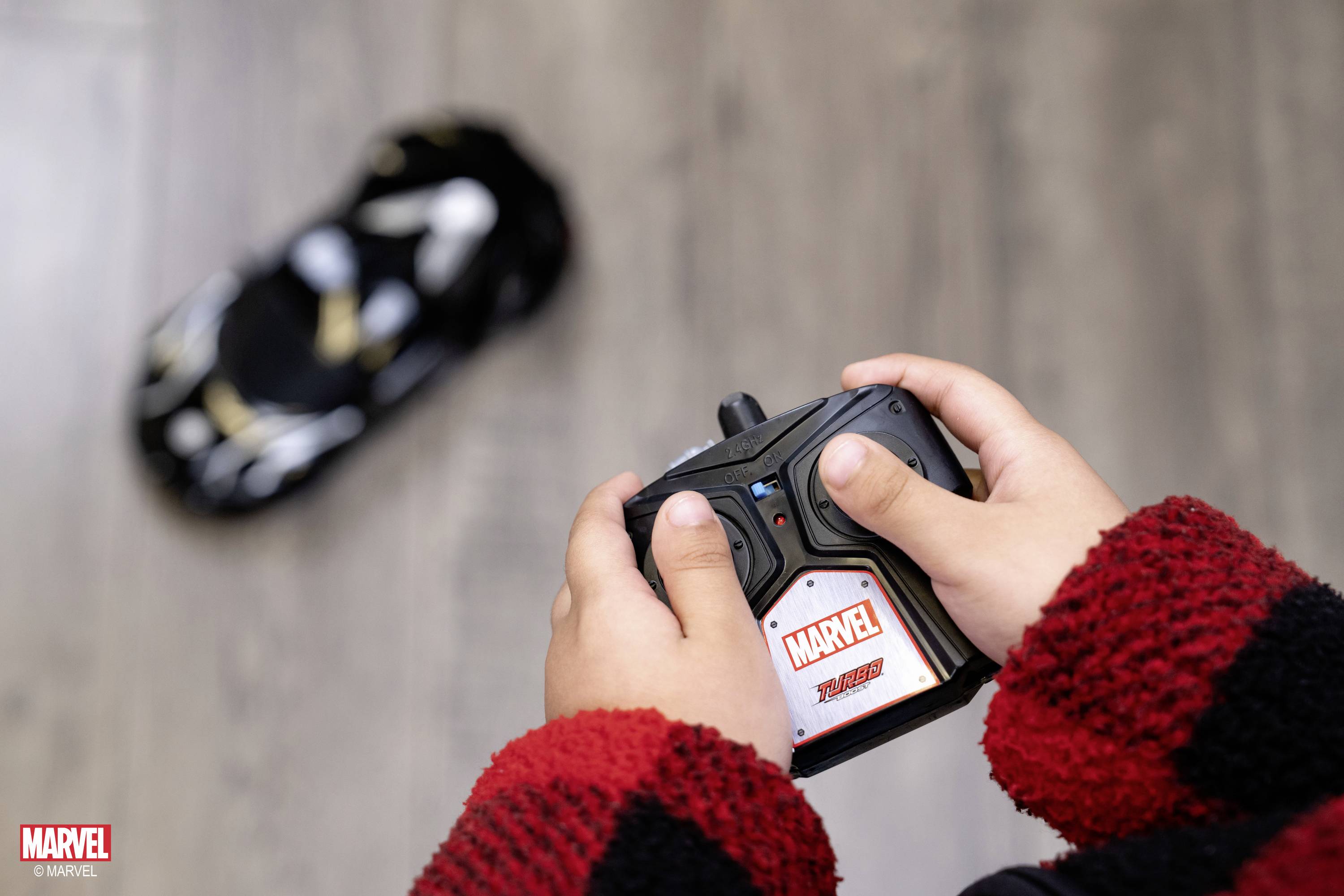 Dziecko steruje samochodem zdalnie sterowanym z pilotem w stylu Marvela. Na tle widać samochód znajdujący się na podłodze.