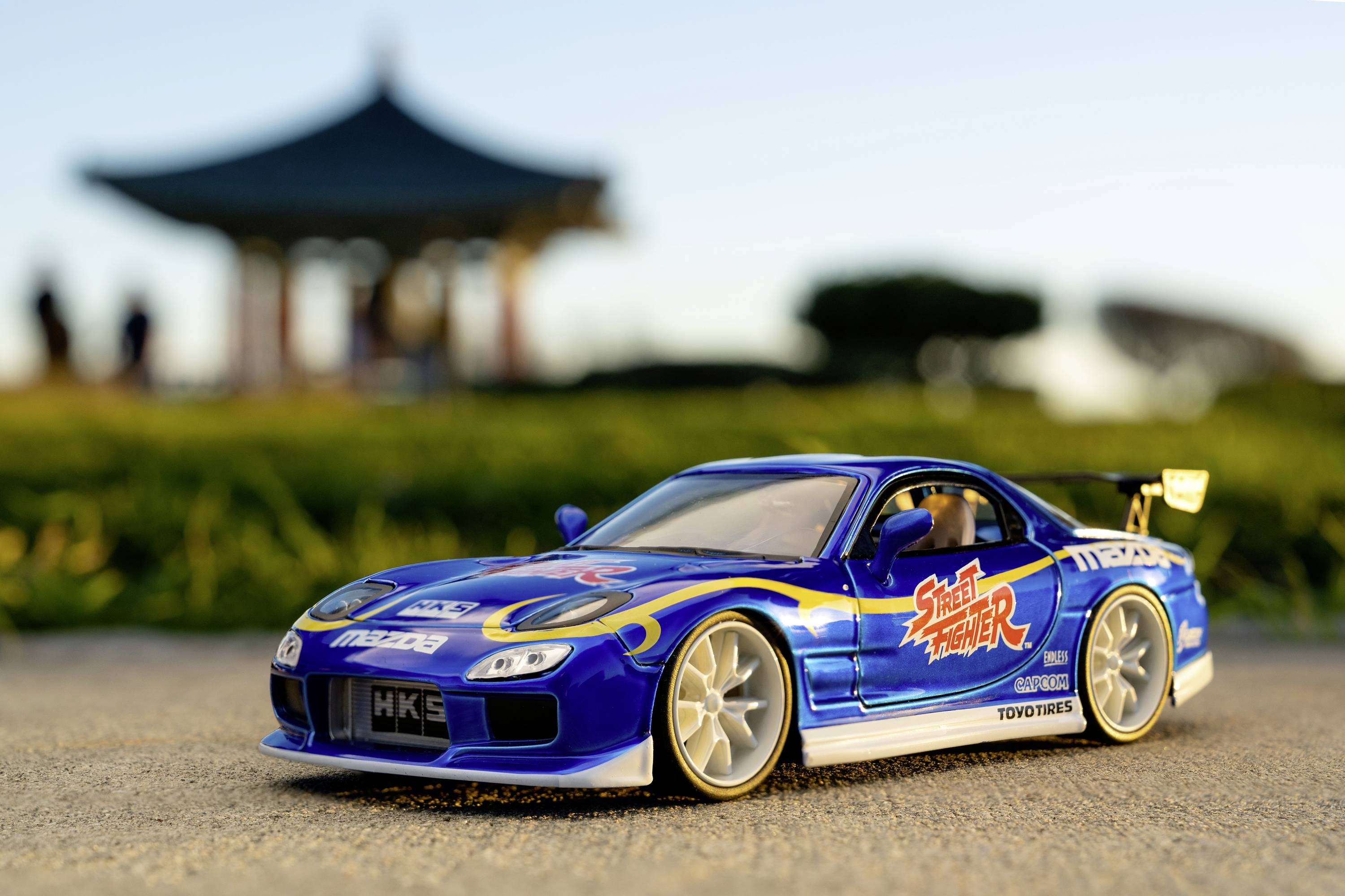 Niebieskie autko modelarskie w stylu wyścigowym na drodze, w tle niewyraźny pawilon. Napis w stylu jaskiniowym na boku samochodu.