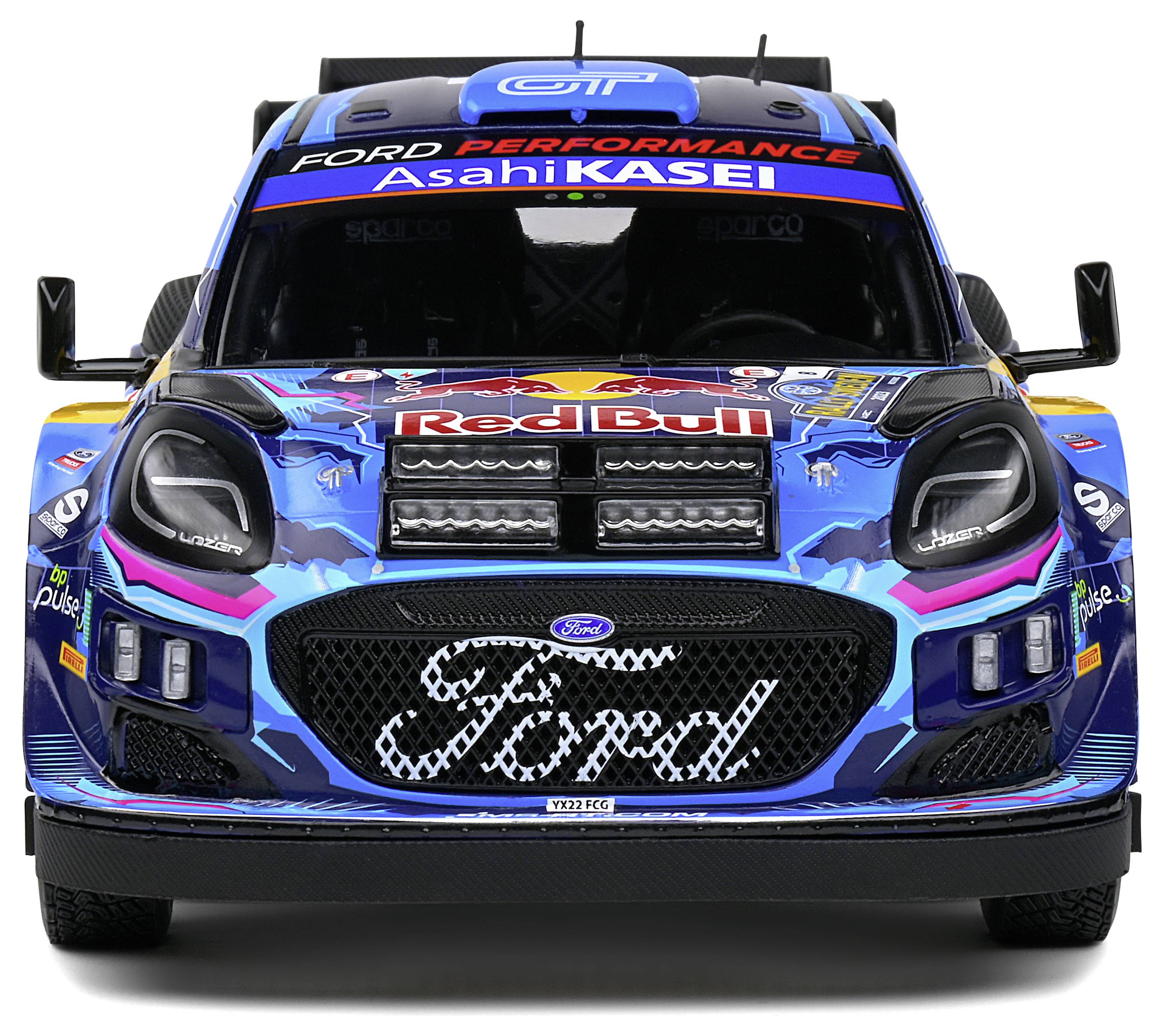 Zdjęcie przedstawia widok z przodu samochodu rajdowego z logo Forda oraz kolorowymi naklejkami sponsorów, w tym 'Red Bull' i 'Asahi KASEI'.