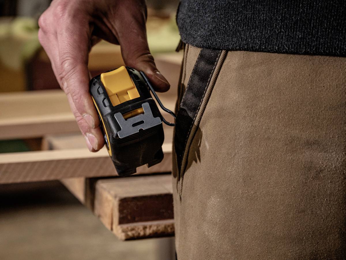 Ręka trzyma miarkę przyłożoną do drewnianej deski. Osoba jest ubrana w brązowe ubranie robocze.