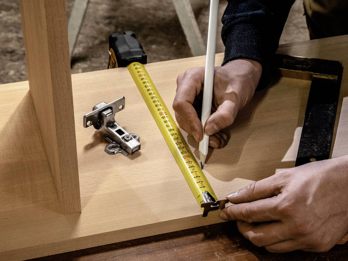 Osoba mierzy drewnianą deskę za pomocą miarki i zaznacza ją ołówkiem. Obok leży zawias.