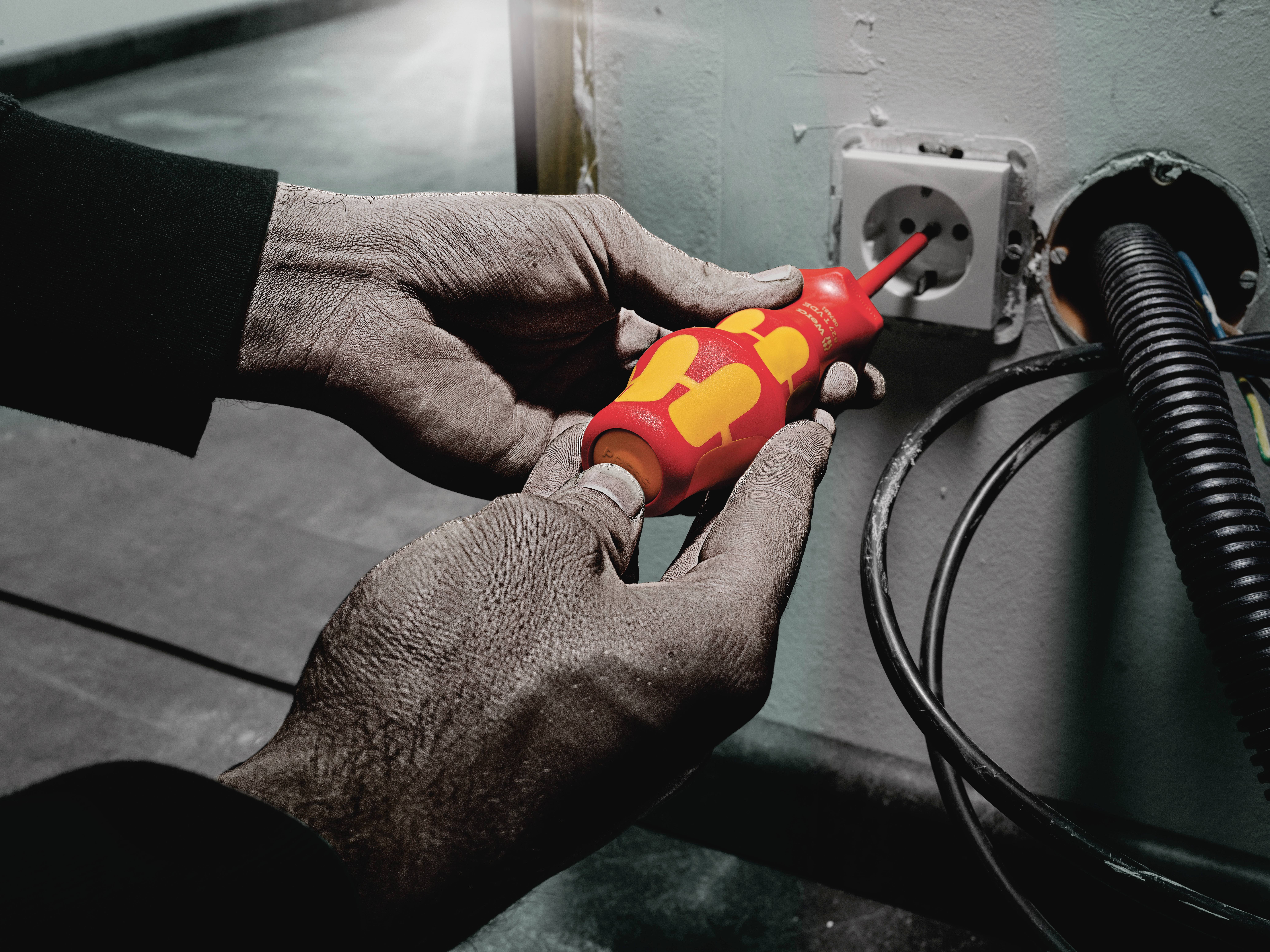 Osoba używa izolowanego śrubokręta do naprawy gniazdka elektrycznego. Przewody są widoczne na ścianie.