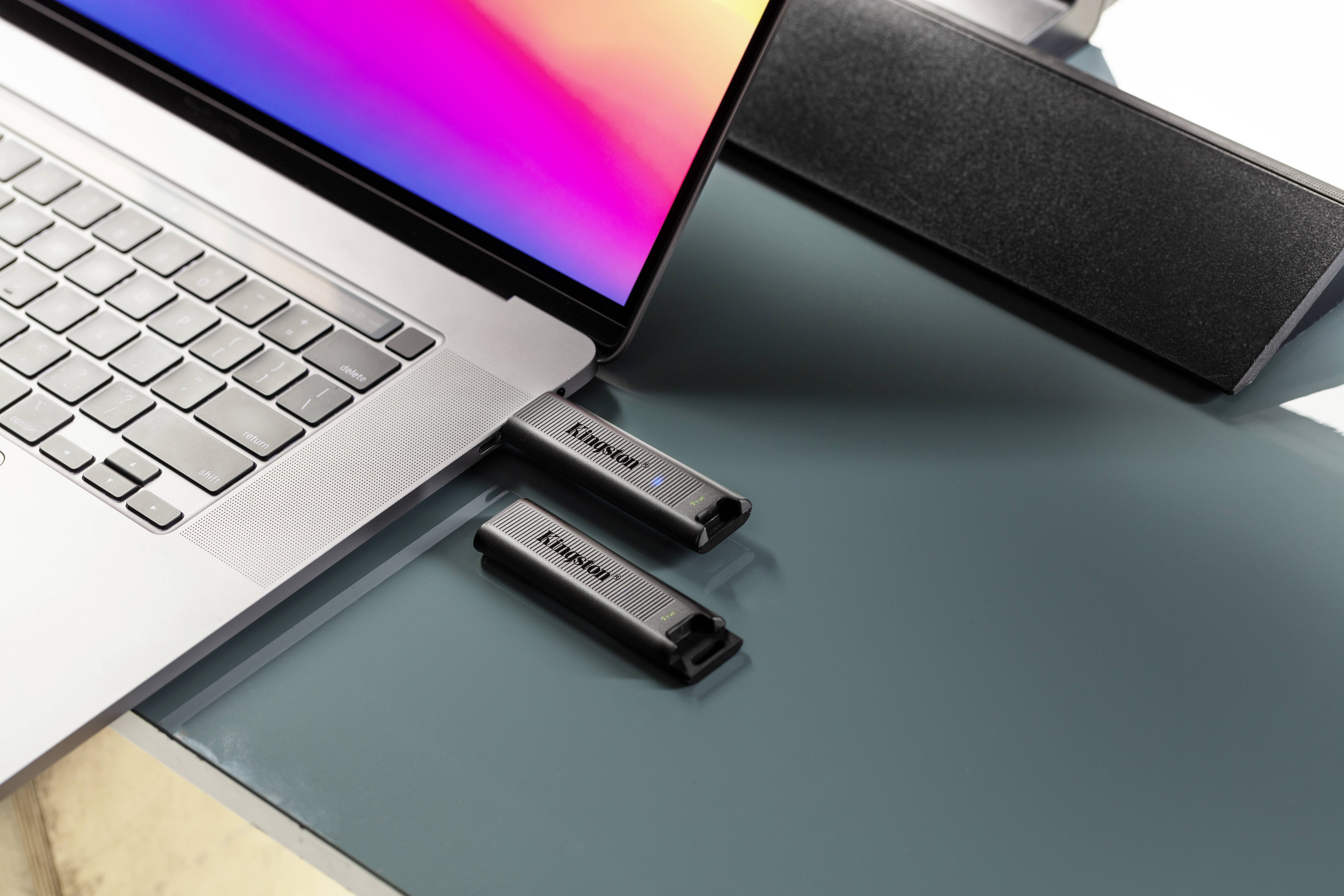 Laptop na stole z podłączonym pendrivem USB i kolejnym leżącym obok. W tle ciemny głośnik.