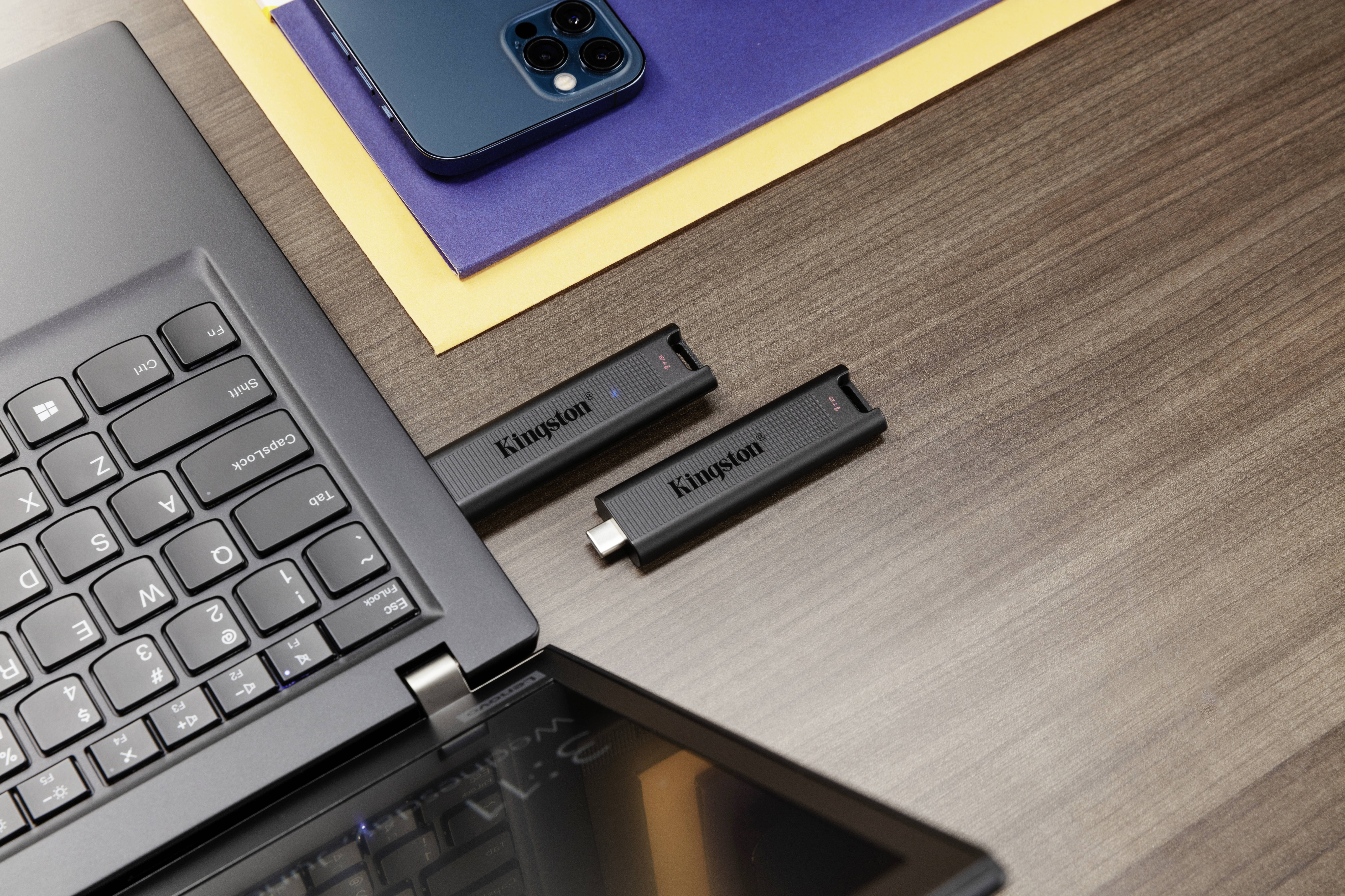 Pendrive'y są włożone do laptopa na stole. Obok laptopa leży smartfon na niebiesko-żółtym papierze.