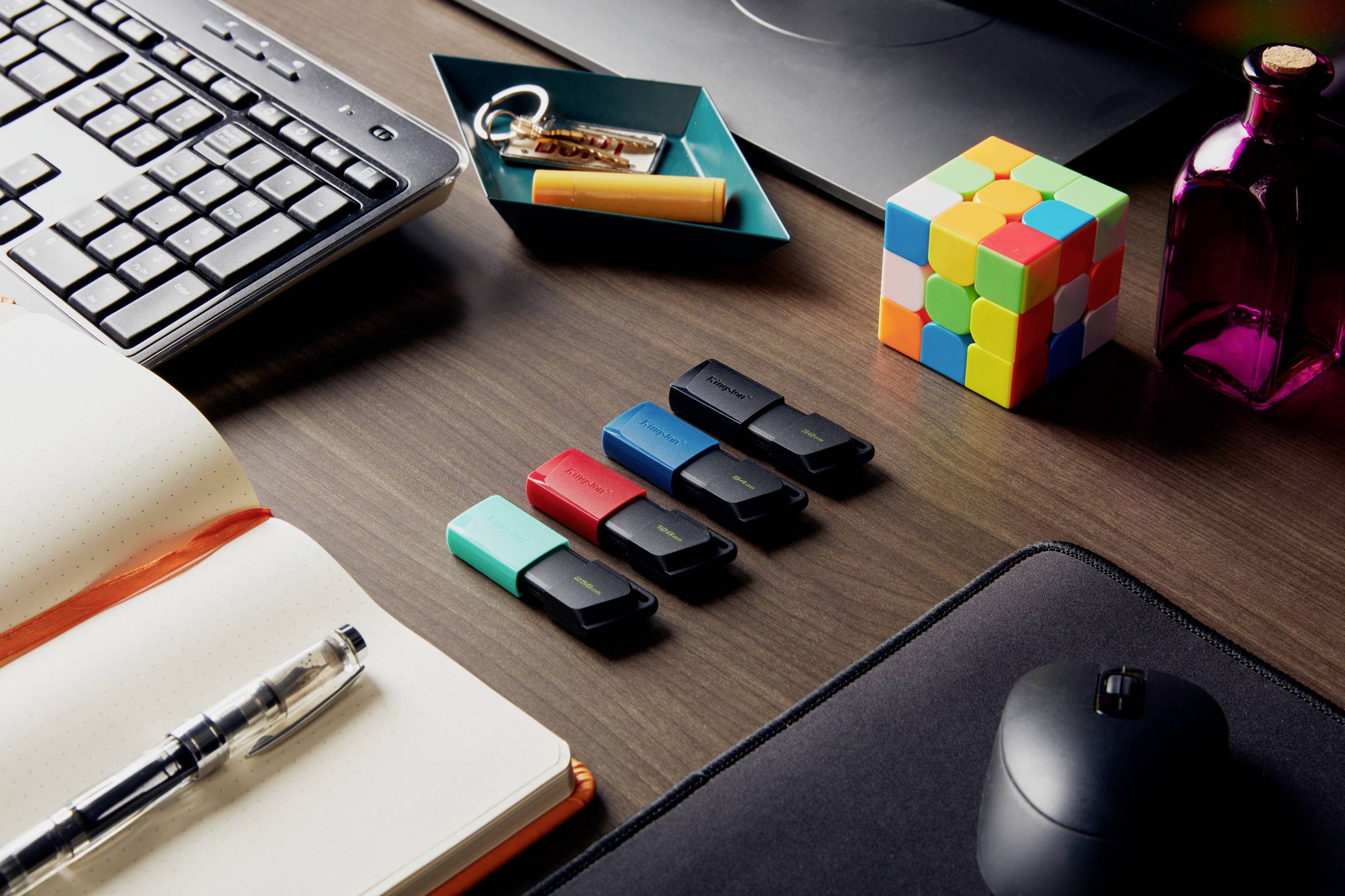 Kilka pendrive'ów w różnych kolorach leży na biurku, otoczonych książką, klawiaturą, kostką i innymi materiałami biurowymi.