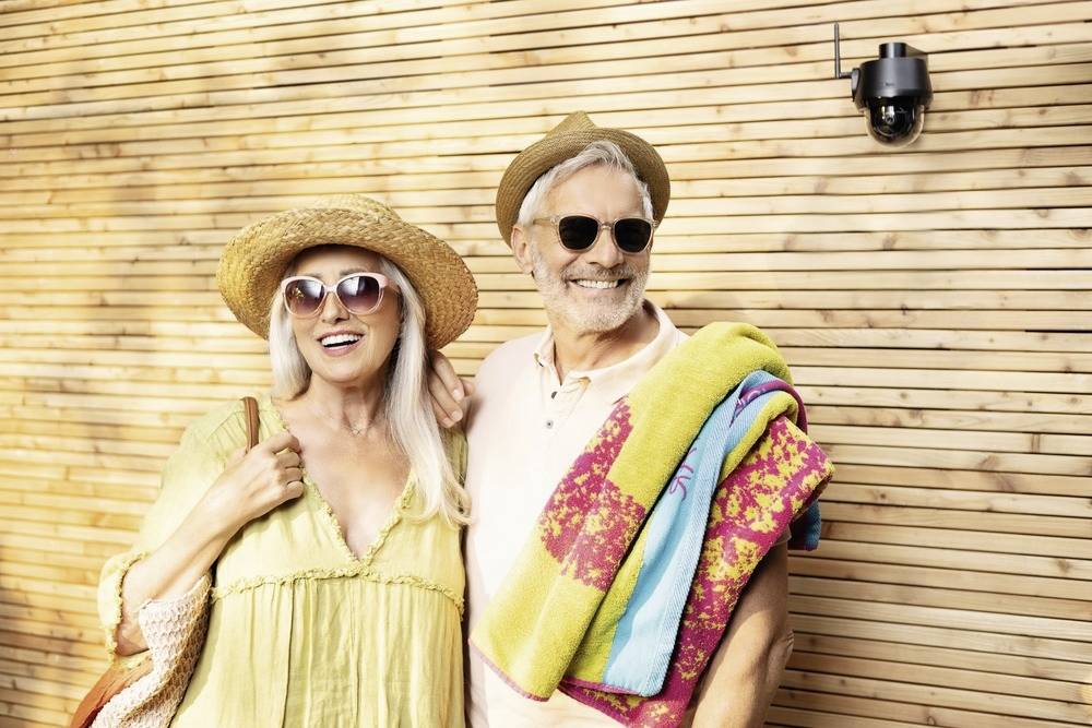 Starsza para w letniej odzieży, w słomianych kapeluszach i okularach przeciwsłonecznych, uśmiecha się przed drewnianą ścianą. Kobieta nosi torbę, a mężczyzna - ręcznik.