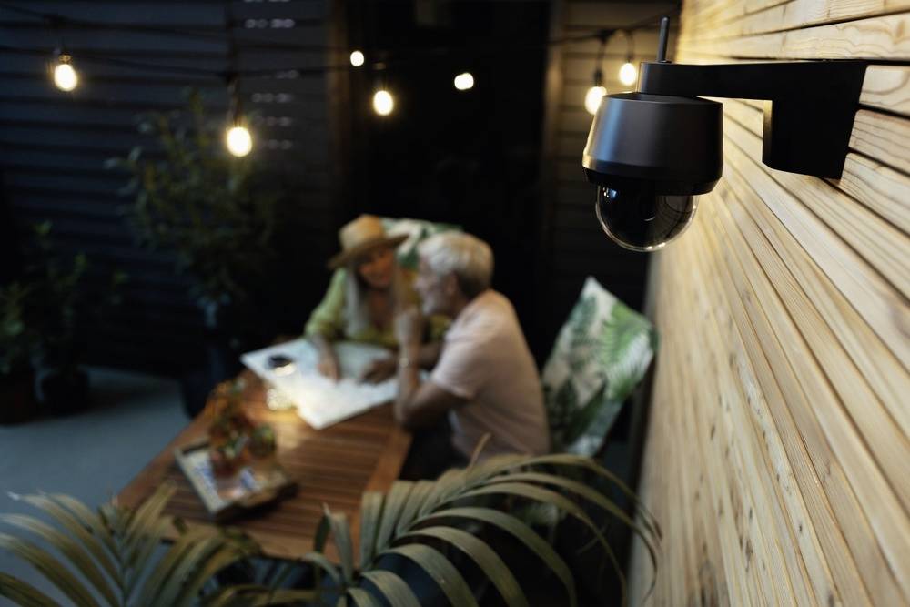 Kamera monitorująca zamocowana na drewnianej ścianie, skierowana na starszą parę siedzącą przy stole i wspólnie studiującą mapę. Panuje zmierzch.