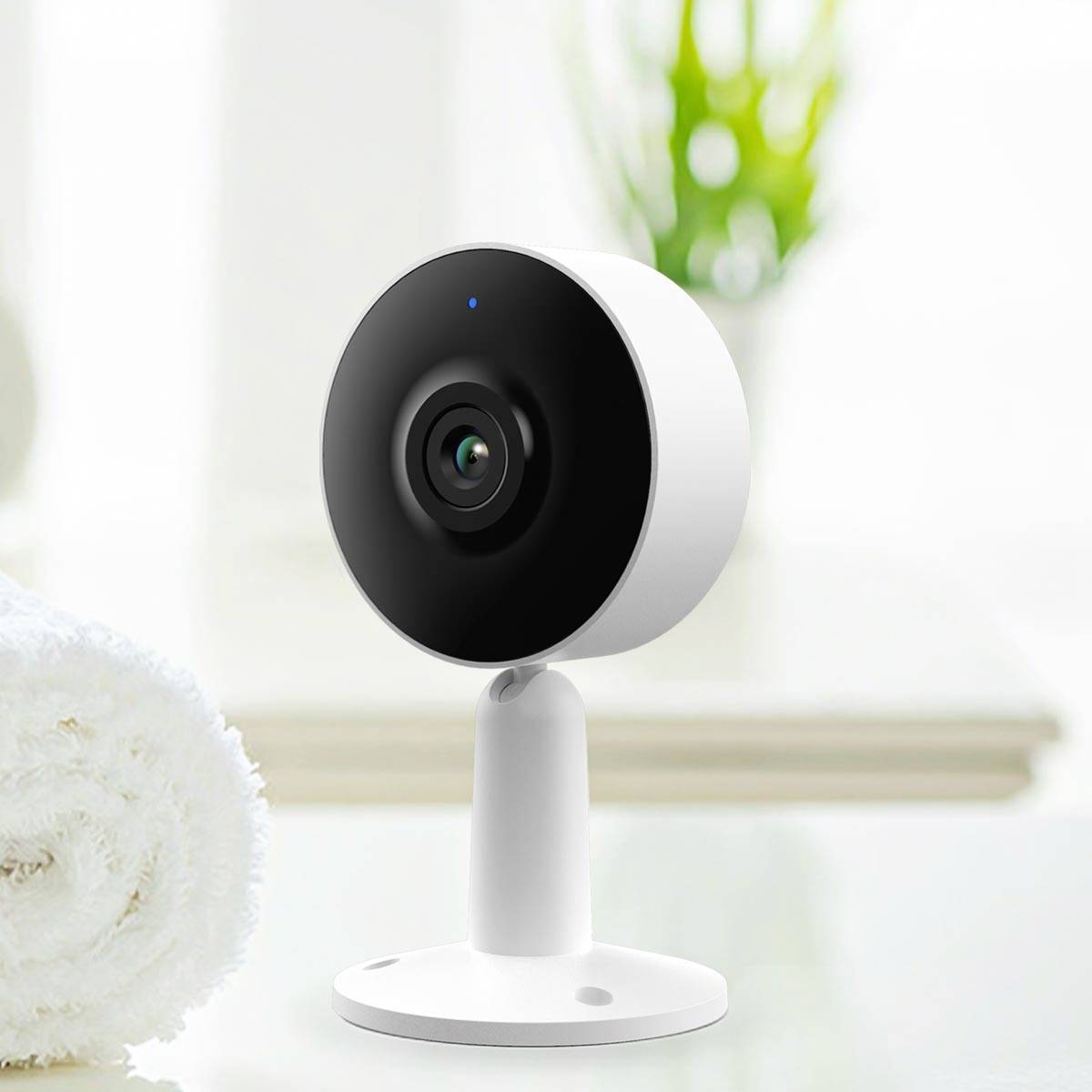 Biała i czarna, okrągła kamera monitoringu stoi na stole obok ręcznika, w tle rozmyte zielone tło.