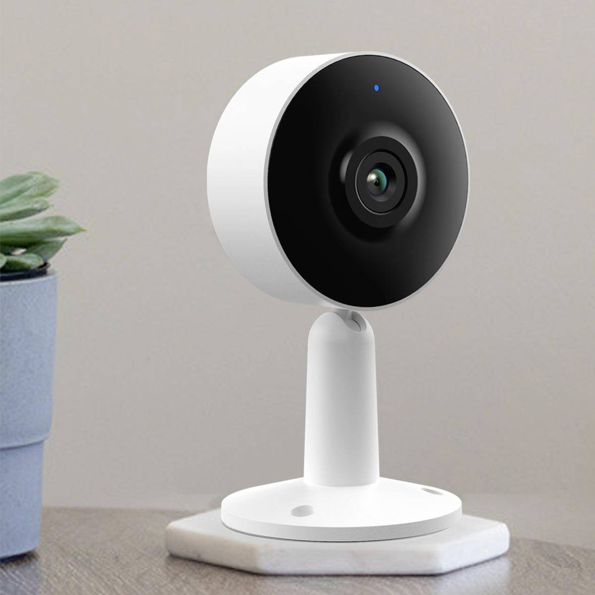 Biała kamera monitoringu umieszczona na podstawce, stojąca na stole. W tle widoczny jest doniczka z rośliną.
