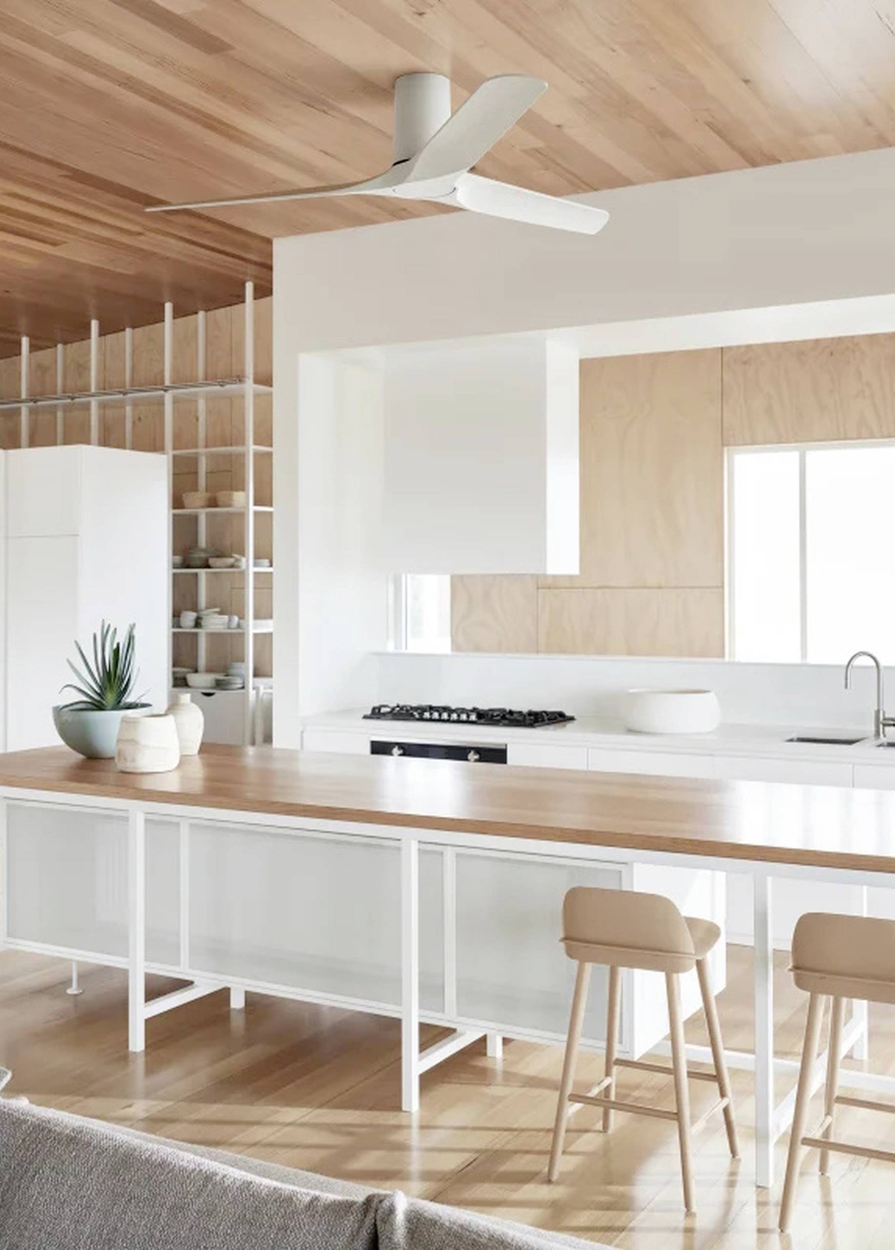 Nowoczesna kuchnia z drewnianym sufitem i wyspą. Białe szafki i hokery barowe, akcent roślinny oraz duże okno tworzą jasną, przestronną atmosferę.