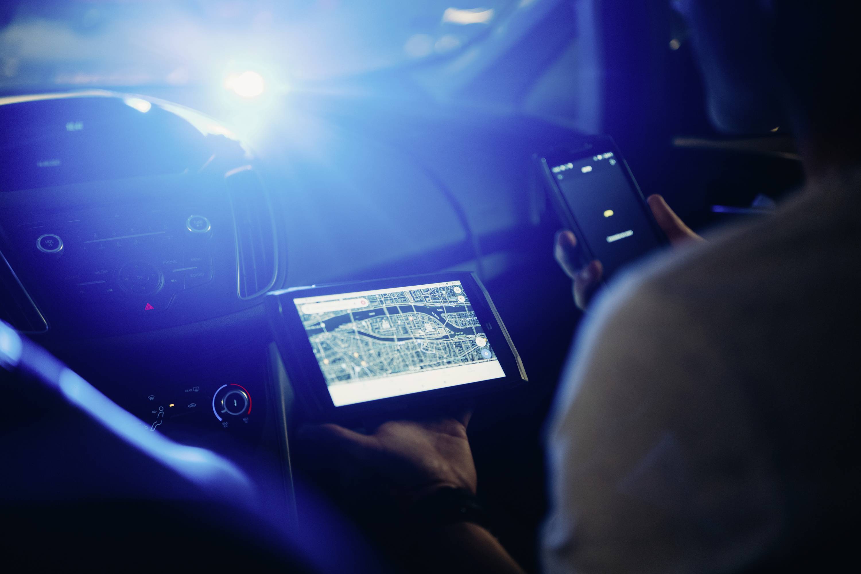 Osoba używa tabletu z mapą miasta w samochodzie, trzymając telefon komórkowy w drugiej ręce, przy świetle nocnym.