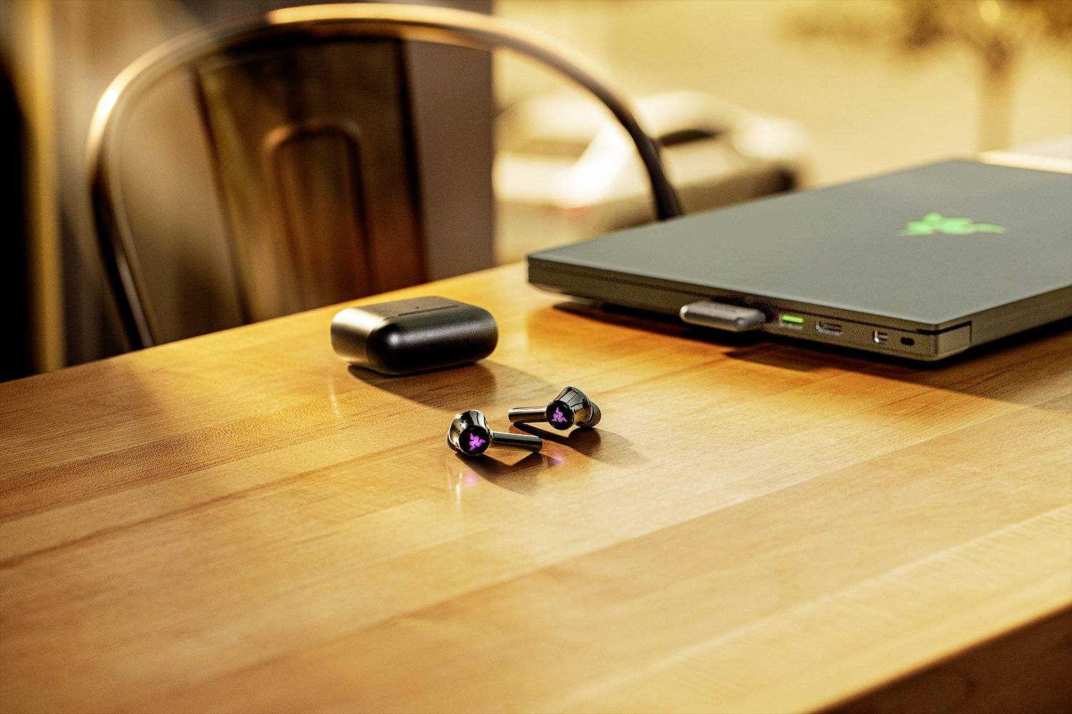 Bezprzewodowe słuchawki douszne i laptop na drewnianym stole. Na zewnątrz świeci słońce, widać rozmyty widok ulicy.