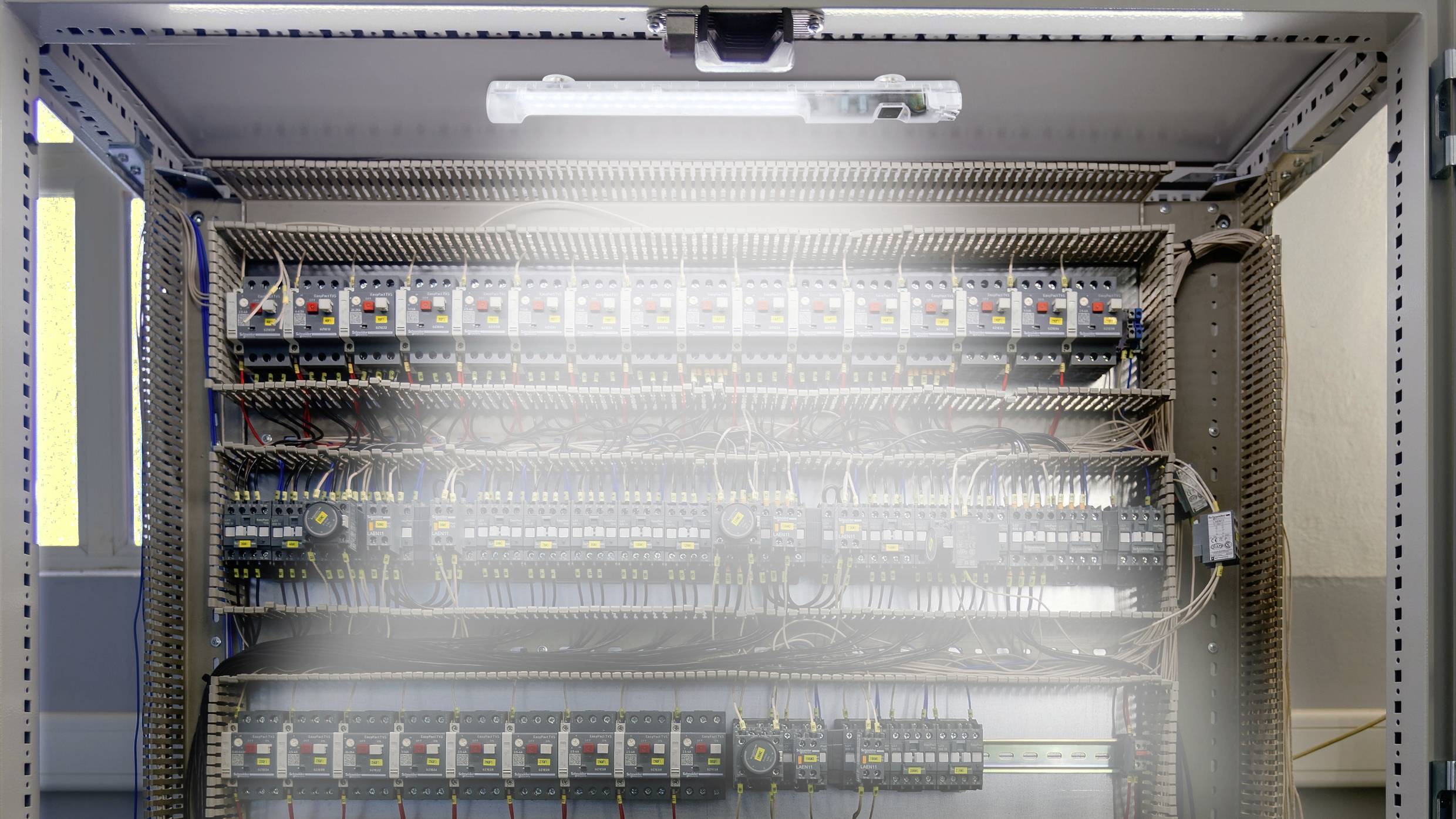 Otwarty rozdzielnik elektryczny z wieloma rzędami bezpieczników i przewodów, oświetlony lampą wewnątrz.