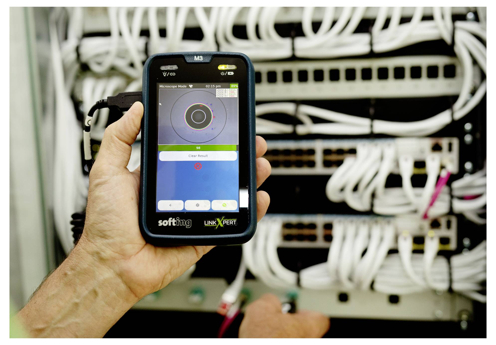 Dłoń trzymająca przenośny tester sieci przed szafą serwerową z wieloma podłączonymi kablami ethernetowymi i przełącznikami.
