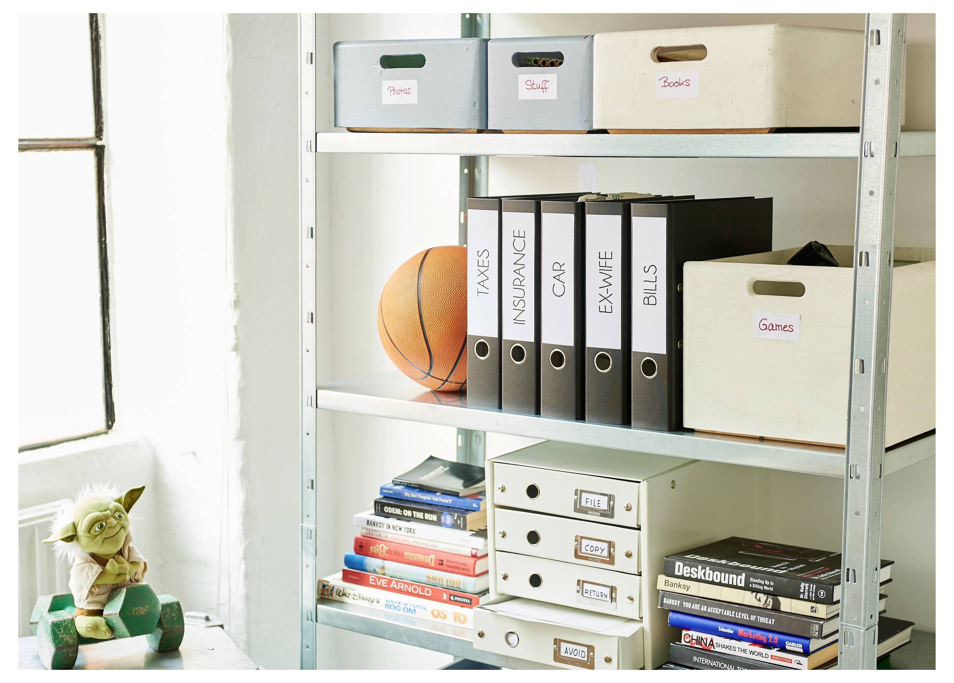 Metalowa półka przechowuje oznaczone pudła do przechowywania, segregatory, książki i piłkę do koszykówki; figurka Yody siedzi przy oknie po lewej stronie.