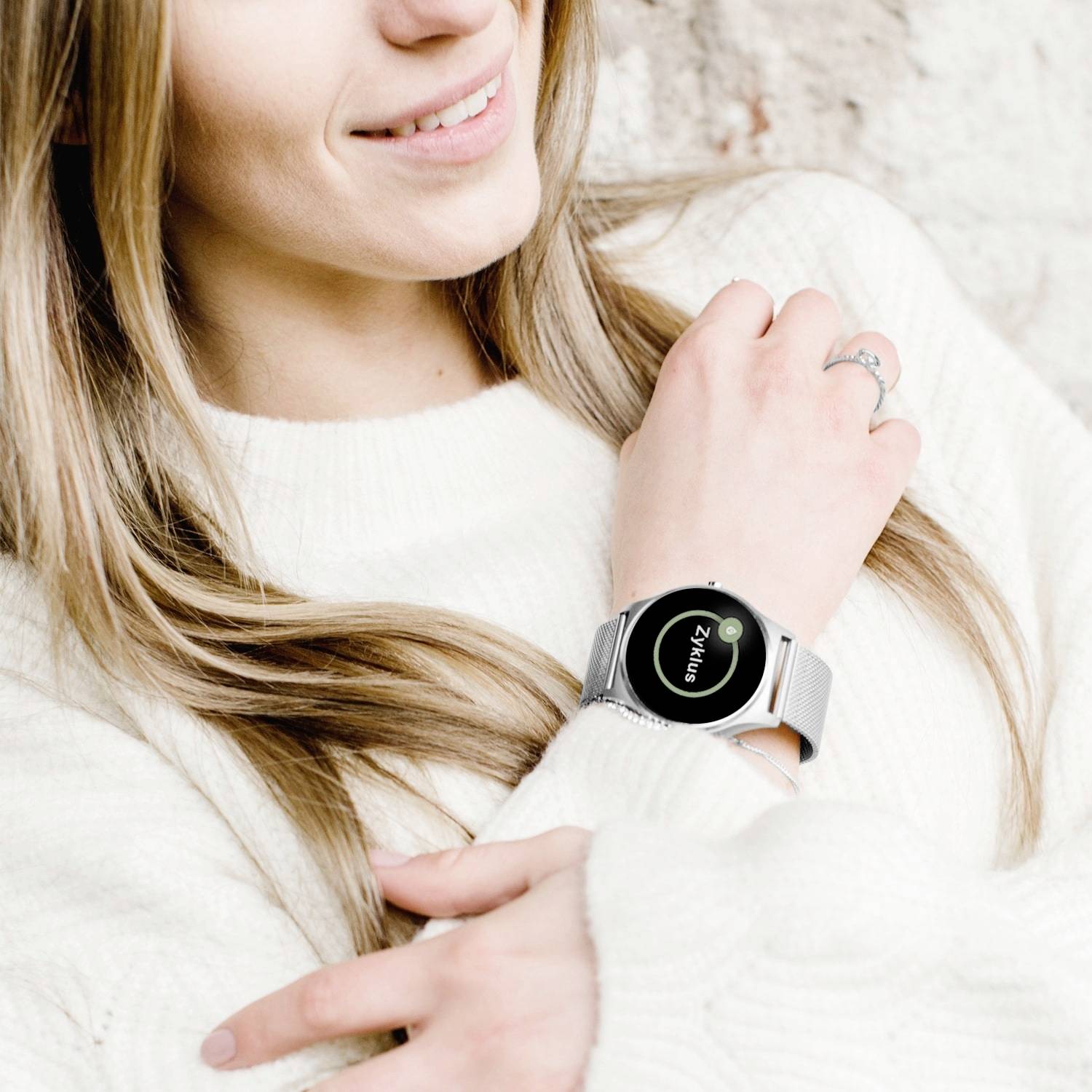 Uśmiechnięta osoba w białym swetrze nosi srebrny smartwatch.