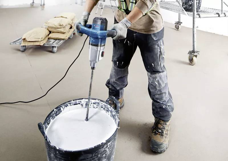 Osoba miesza białą farbę elektrycznym mieszadłem w dużym wiadrze. W tle leżą worki na platformie.