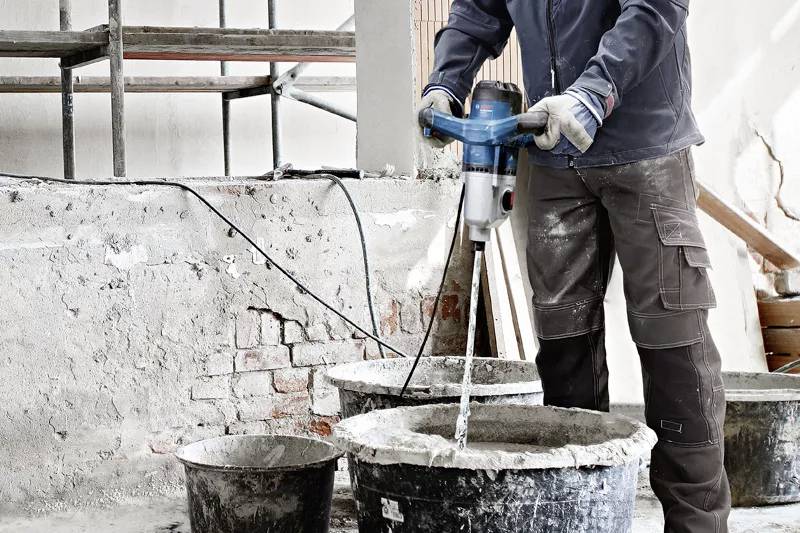 Robotnik budowlany miesza cement elektrycznym mieszadłem w wiadrze. W tle widoczne są ściany i rusztowanie.