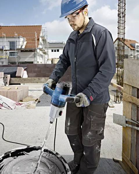 Robotnik budowlany miesza cement na placu budowy za pomocą elektrycznego mieszadła ręcznego. W tle widoczne są rusztowania i stosy cegieł.
