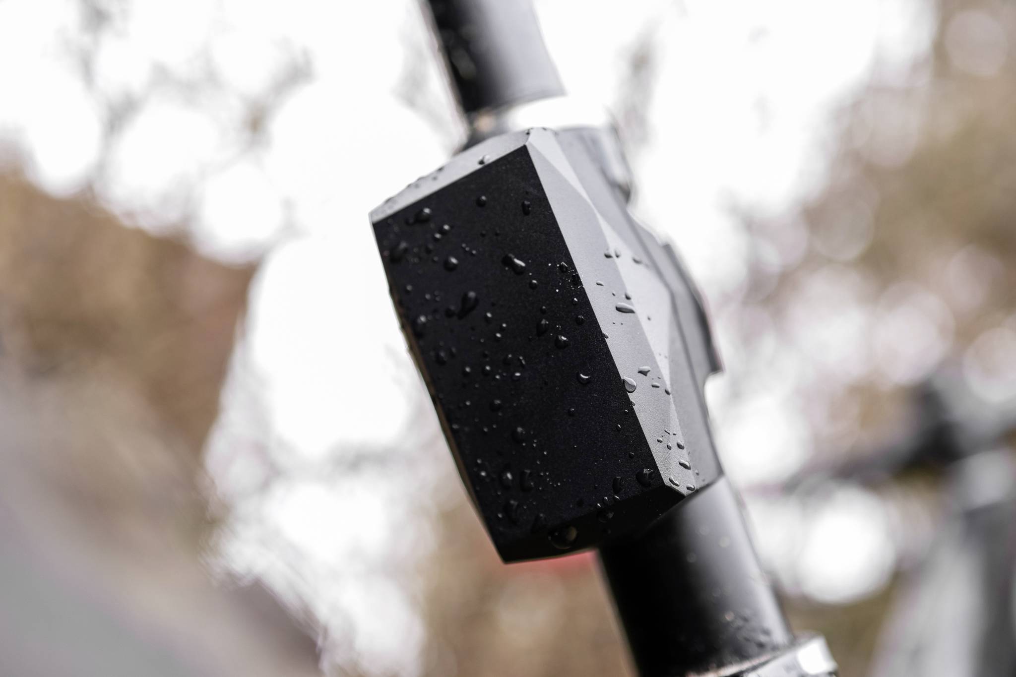 Czarne urządzenie pokryte kropelkami wody jest zamocowane na rurze lub kierownicy. W tle widać nieostre gałęzie drzew.