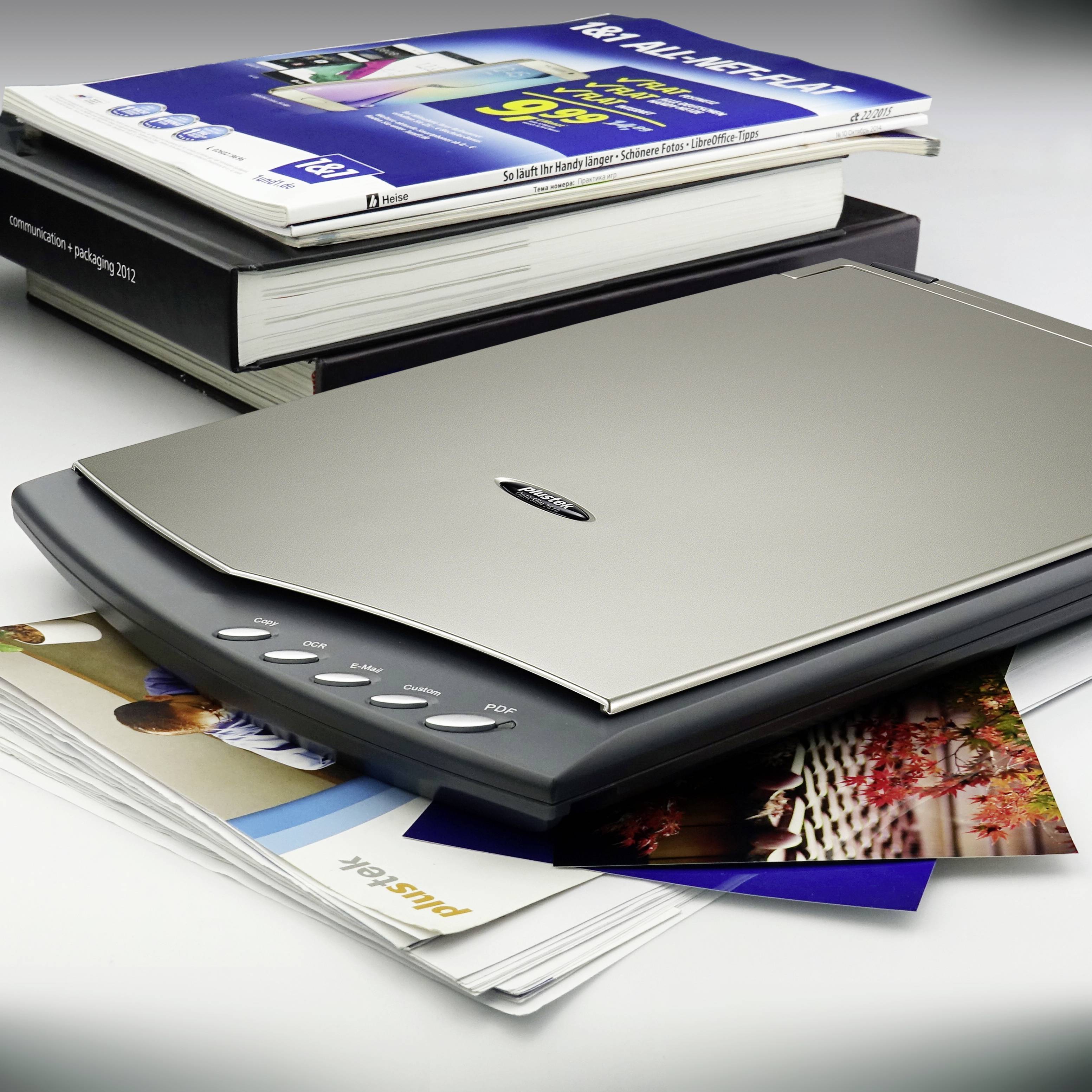 Skaner płaski leży na stosie książek i czasopism. Skaner posiada przyciski do obsługi funkcji. W tle widoczne są książki.