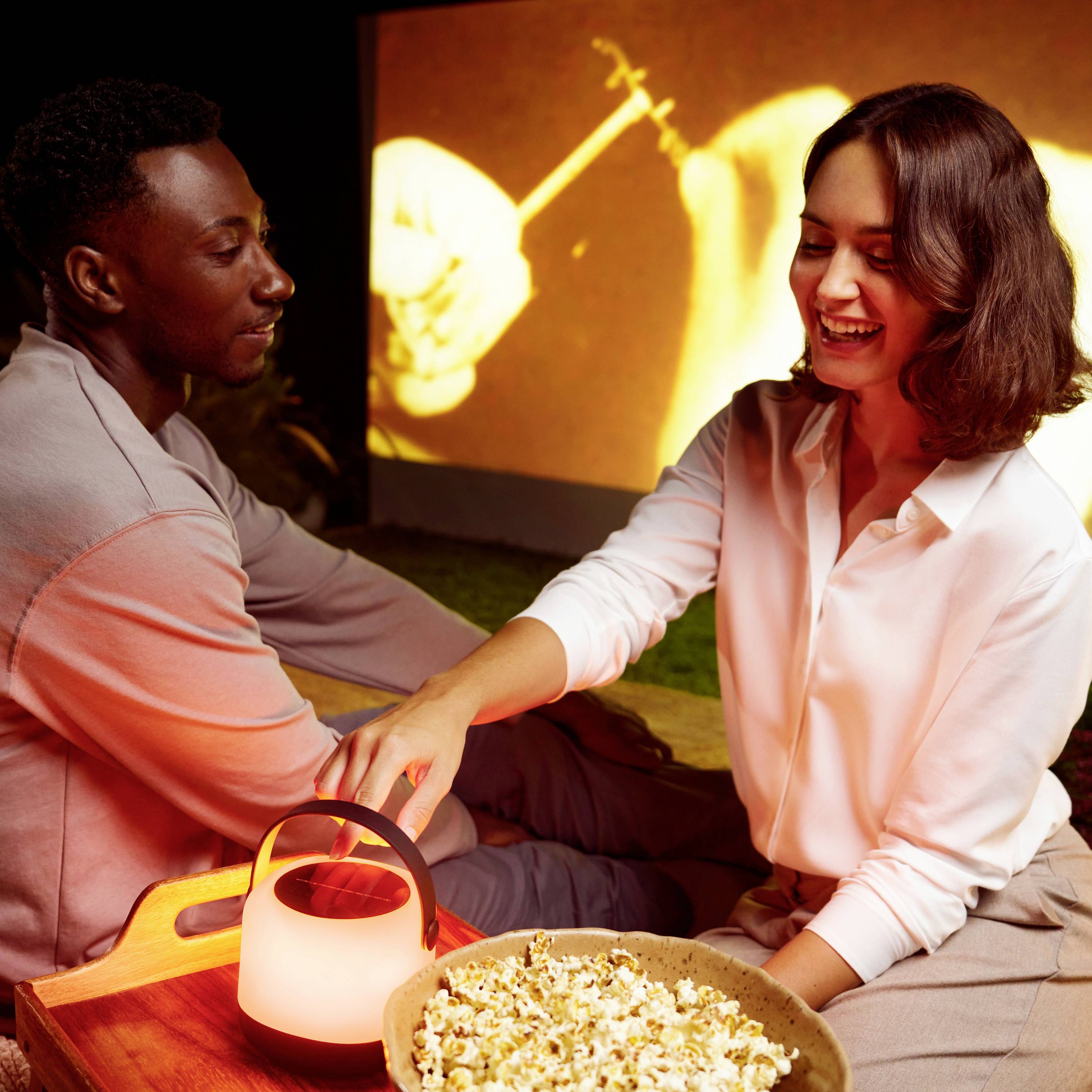 Mężczyzna i kobieta siedzą uśmiechnięci z misą popcornu przed projektorem wyświetlającym film, w przytulnej atmosferze.