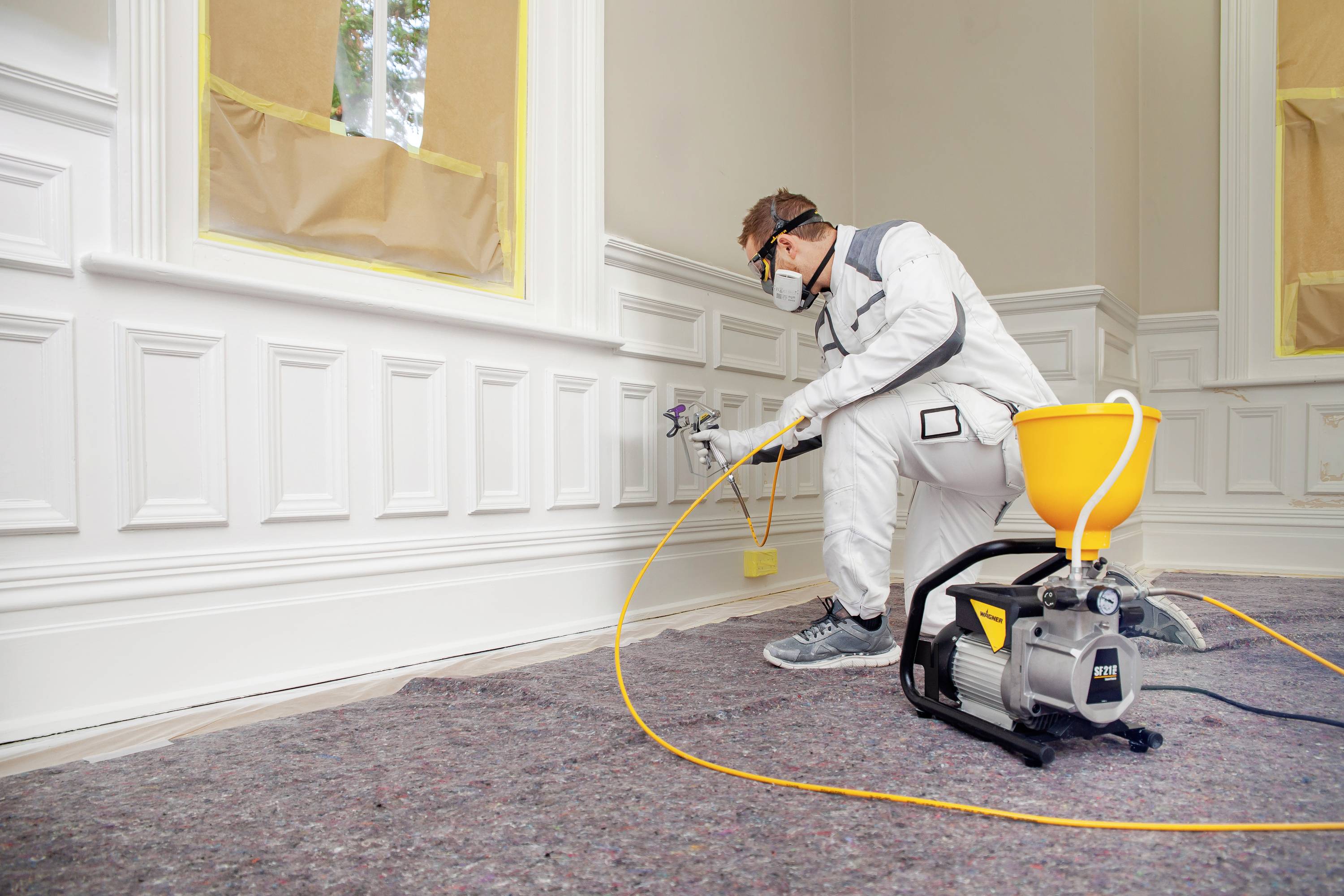 Malarz rozpyla białą farbę ze spryskiwacza na ścianę w odremontowanym pomieszczeniu. Okna są zabezpieczone taśmą malarską.