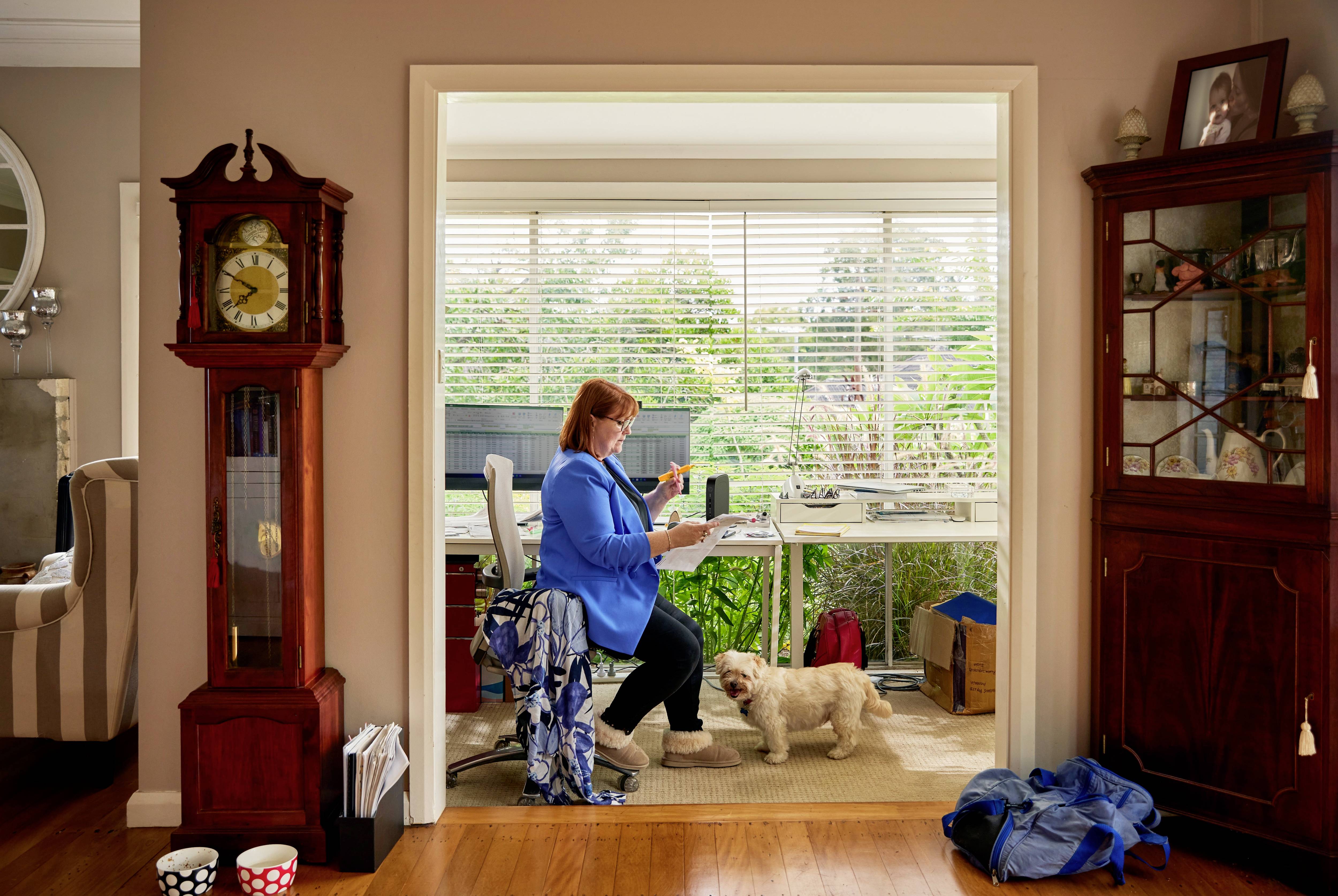 Osoba w niebieskiej marynarce pracuje przy biurku w domu, a mały pies siedzi obok niej. Jasne, przyjazne otoczenie.