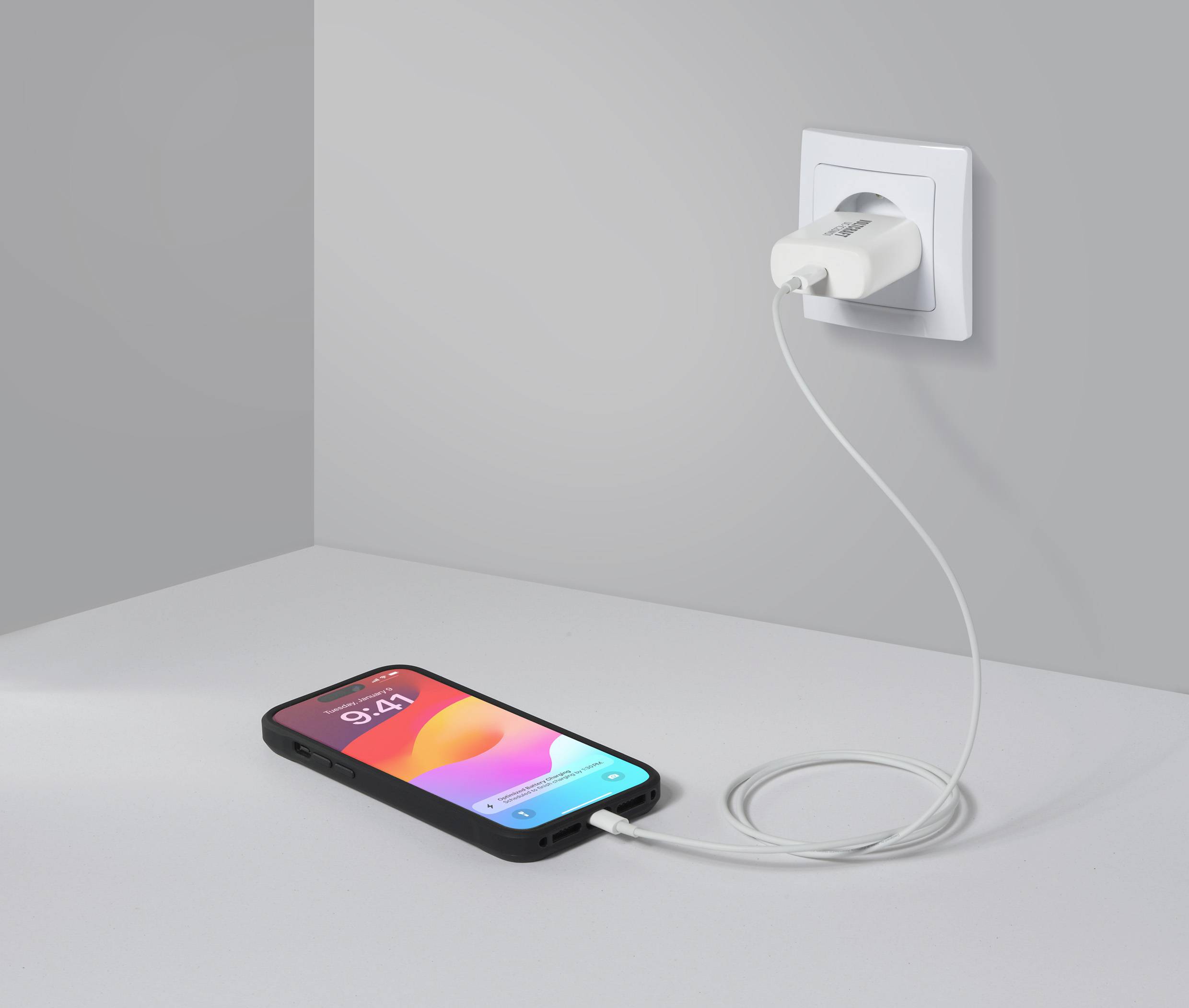 Smartfon ładuje się przez kabel podłączony do gniazdka elektrycznego. Wyświetlacz pokazuje godzinę 9:41 oraz kolorową grafikę w tle.