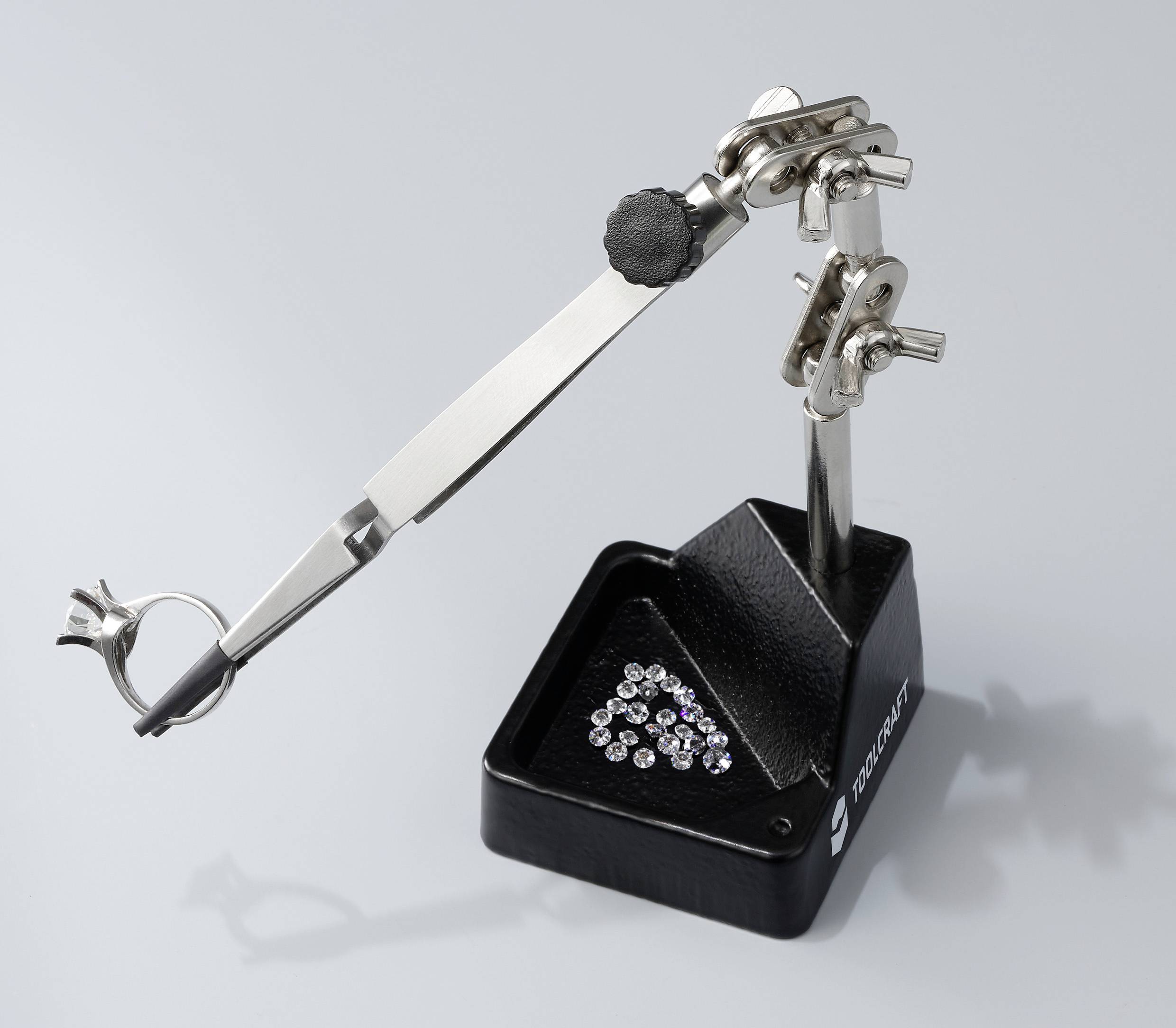 Przyrząd mocujący z metalowymi zaciskami i lupą, używany do precyzyjnych prac, takich jak wytwarzanie biżuterii. Poniżej leżą małe diamenty.