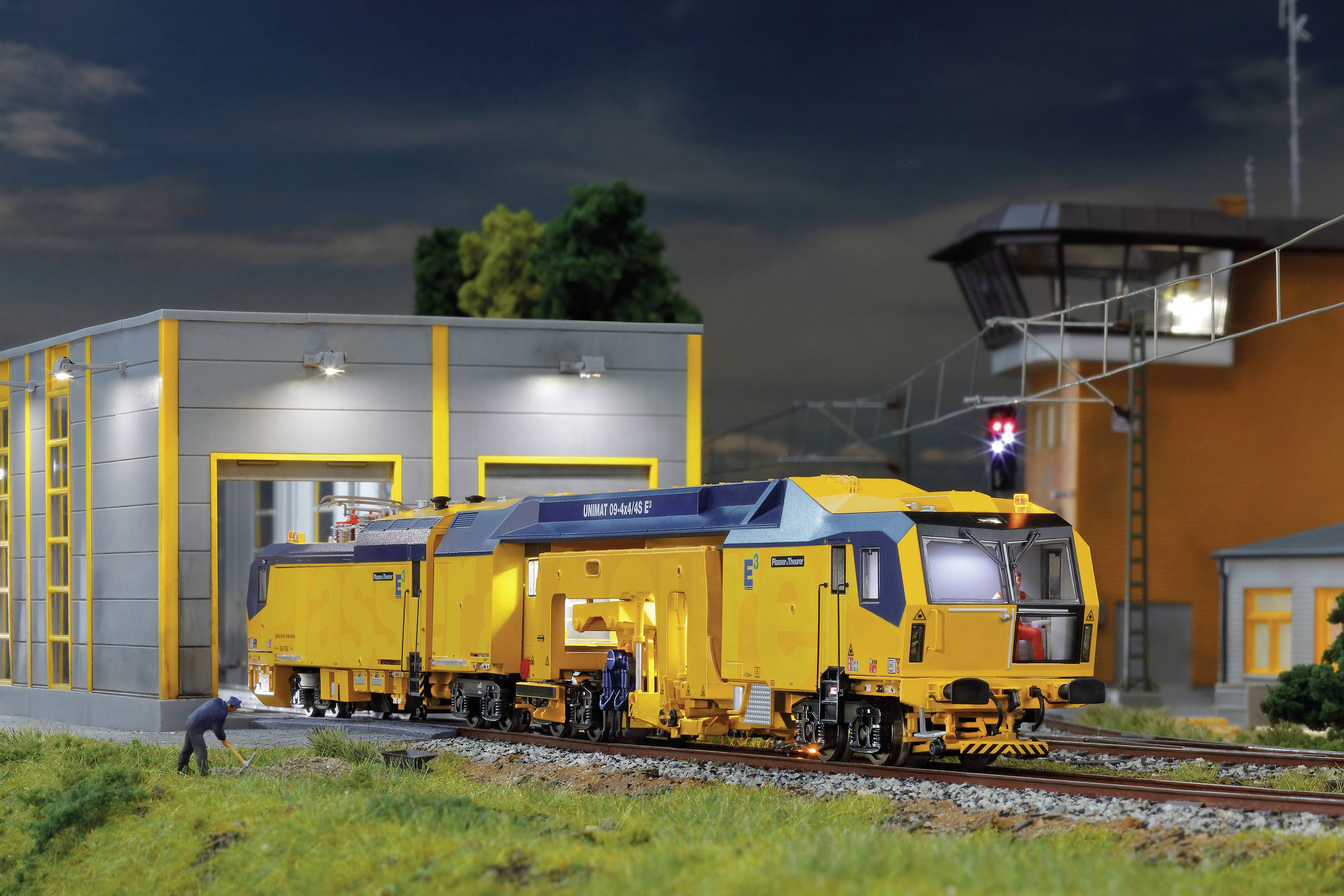 Żółta lokomotywa stoi na torach przed oświetlonym budynkiem. Na pierwszym planie dwóch robotników pracuje przy lokomotywie.